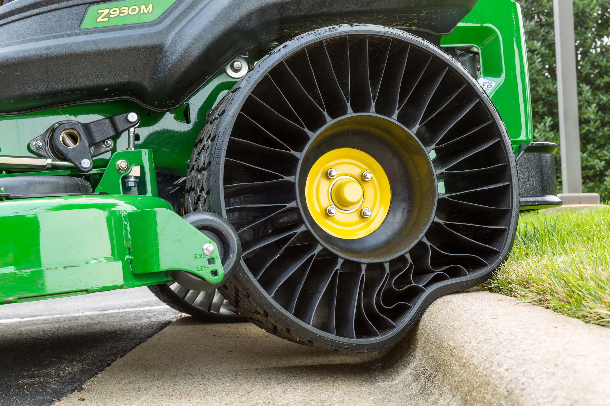 Close-up of a Michelin X Tweel airless rear tire on a mower as it rolls over a curb, showing the flexible spoke design compressing against the concrete edge