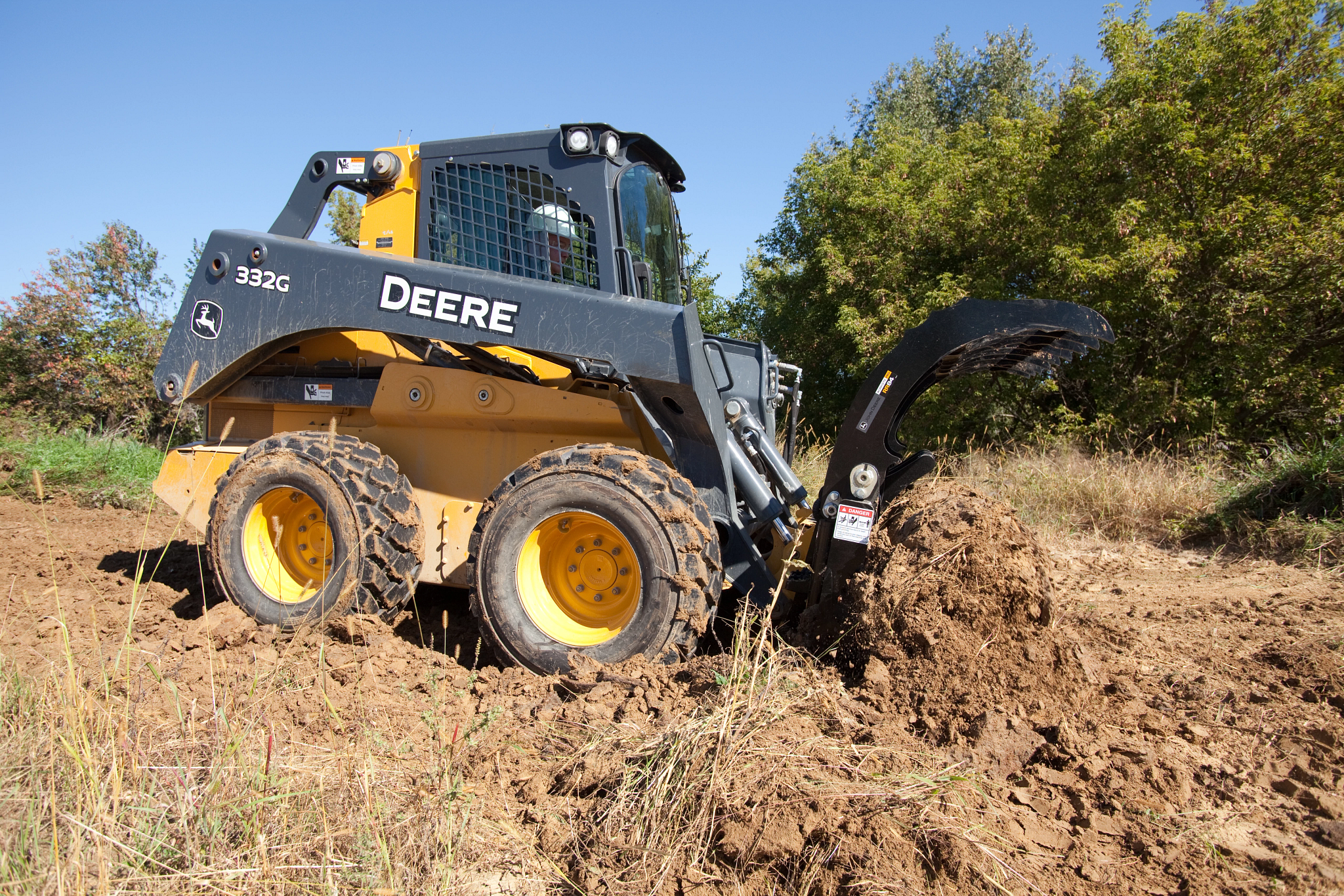 Side right view of a 332G Skid Steer with RR84 Root Rake jaws open wide on a mound of dirt at a dirt-and-forested jobsite.