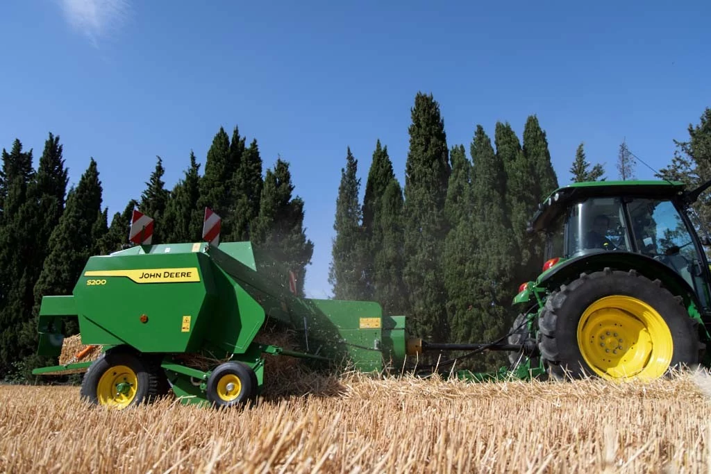 Green tractor pulling a John Deere S200 square baler through a dry field of cut straw, producing rectangular hay bales. Tall trees line the background under a clear blue sky, with straw scattered across the ground.