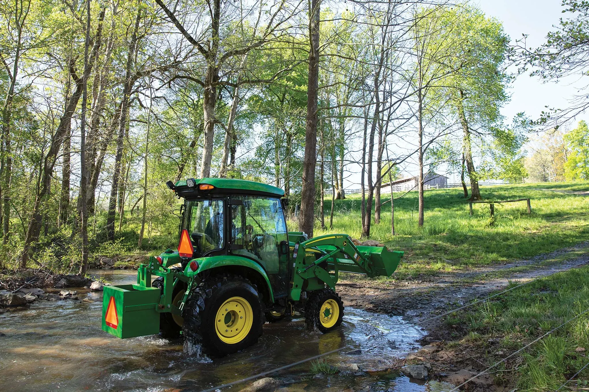 Side right view of a 3046R compact utility tractor crossing a stream in a wooded area