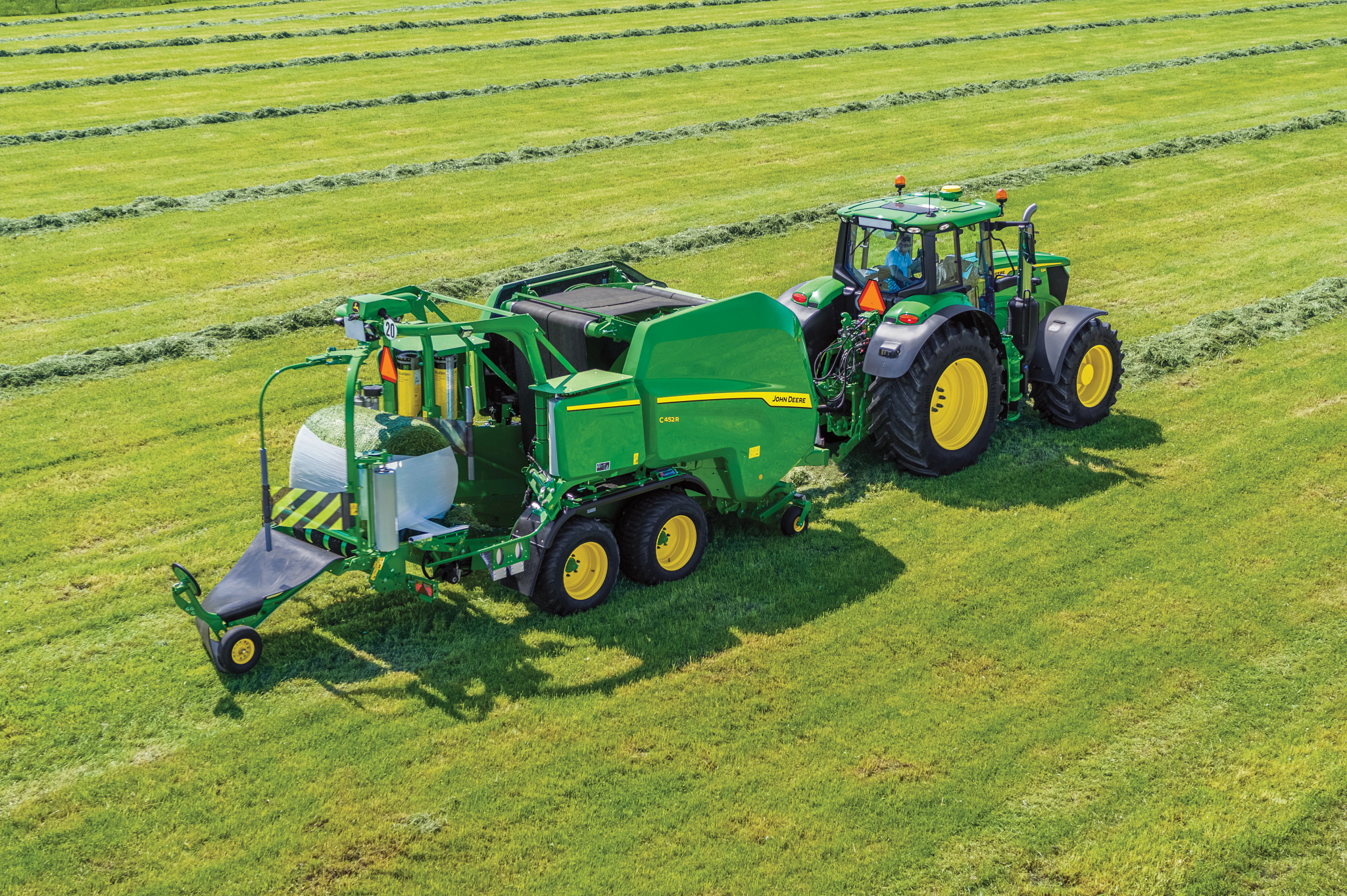 Aerial view of John Deere tractor pulling a green round baler with integrated bale wrapper, forming wrapped hay bales in a cut grass field with parallel windrows.