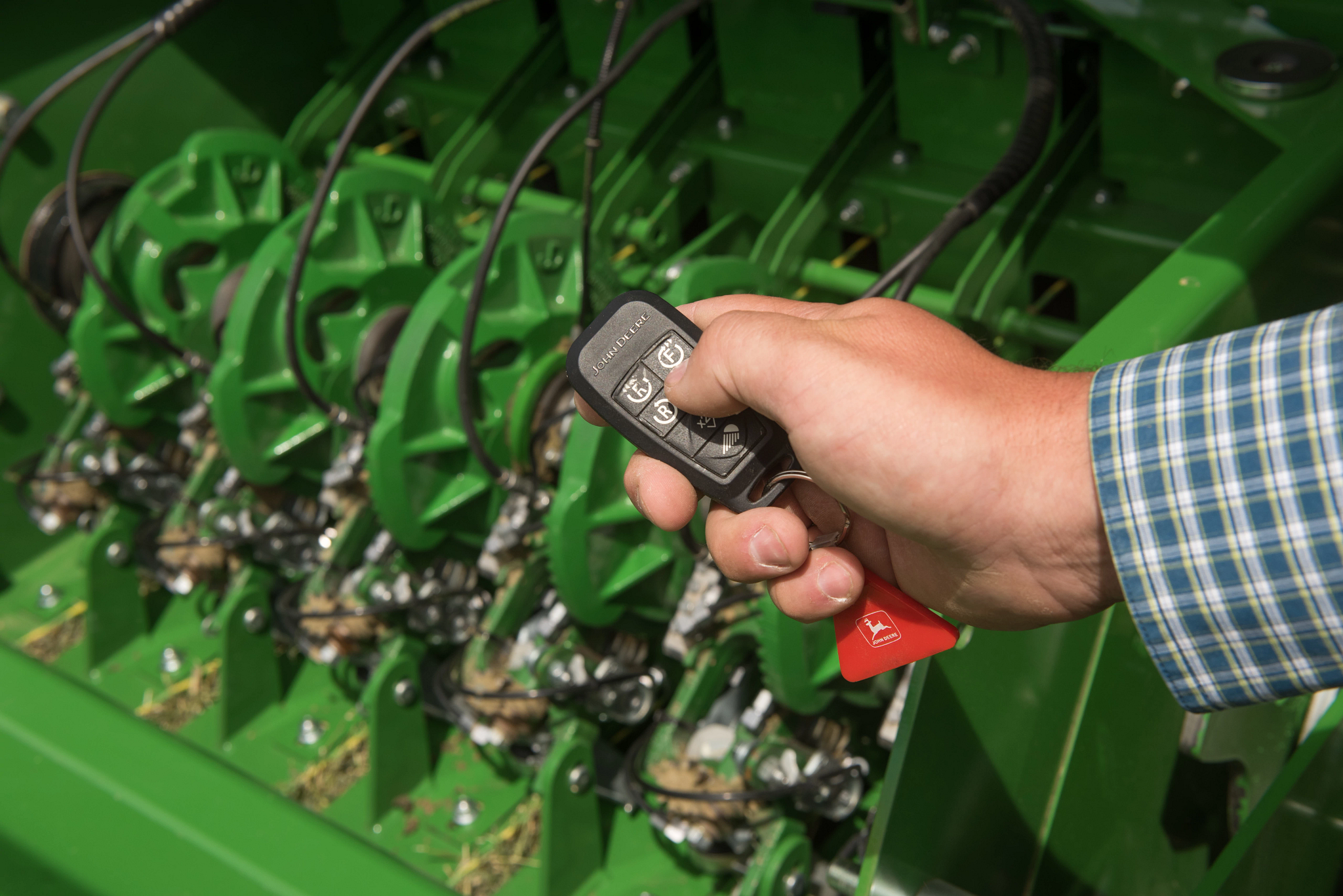 A close-up of a hand holding a small remote above green mechanical baler assemblies with connected cables and metal parts