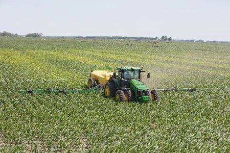 A tractor pulling a liquid applicator moves through a large, green crop field with the sprayer’s booms extend widely on both sides under a bright, clear sky