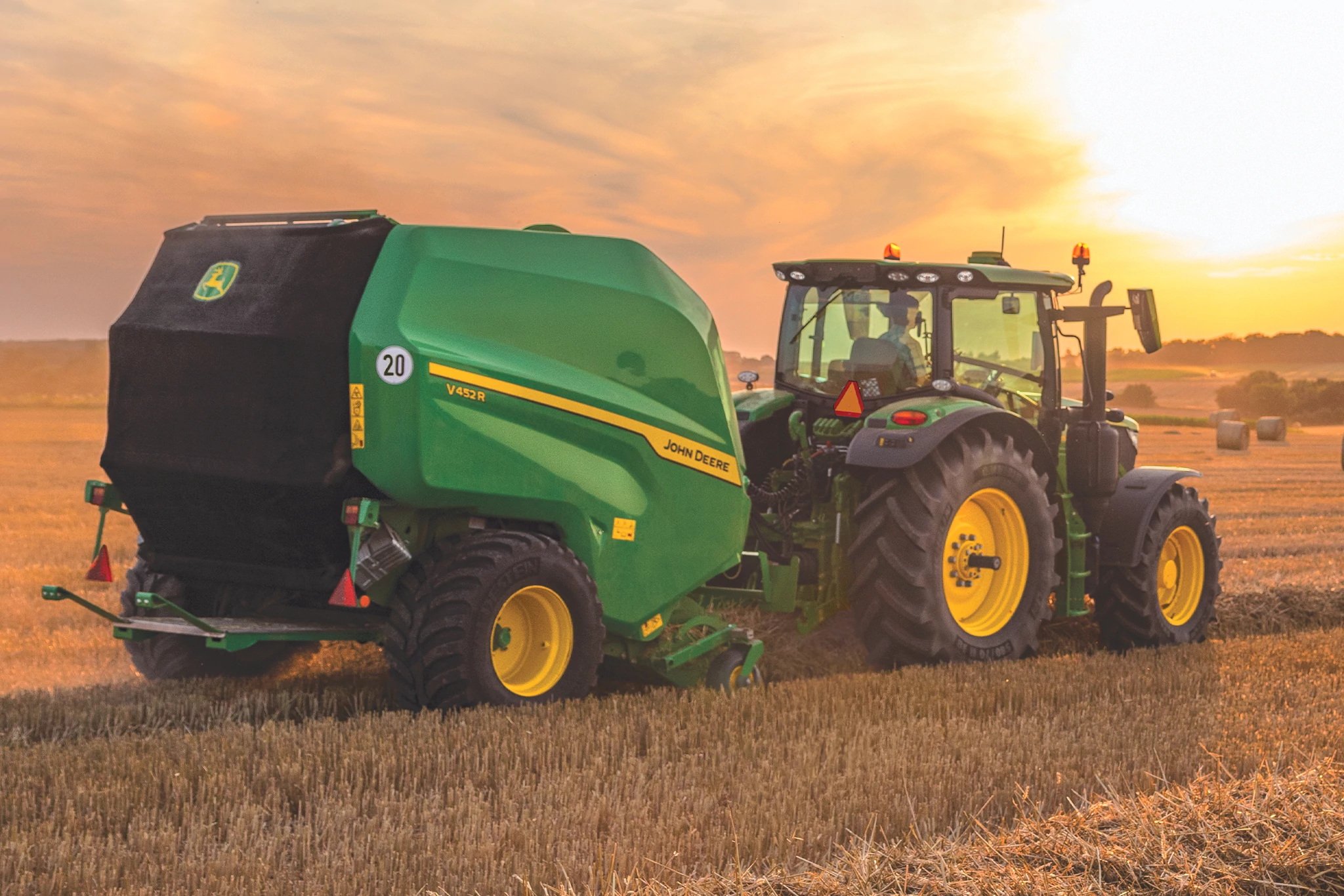 Rear right view of a 6R tractor pulling a V452R round baler across a golden field at sunset