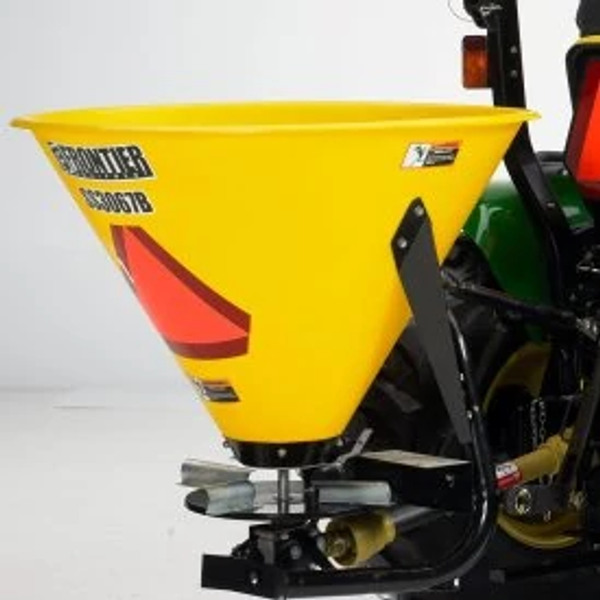 Close up, front left studio view of a yellow SS3067B broadcast spreader with orange high-vis triangle pasted to the side. Part of the attached tractor is visible.