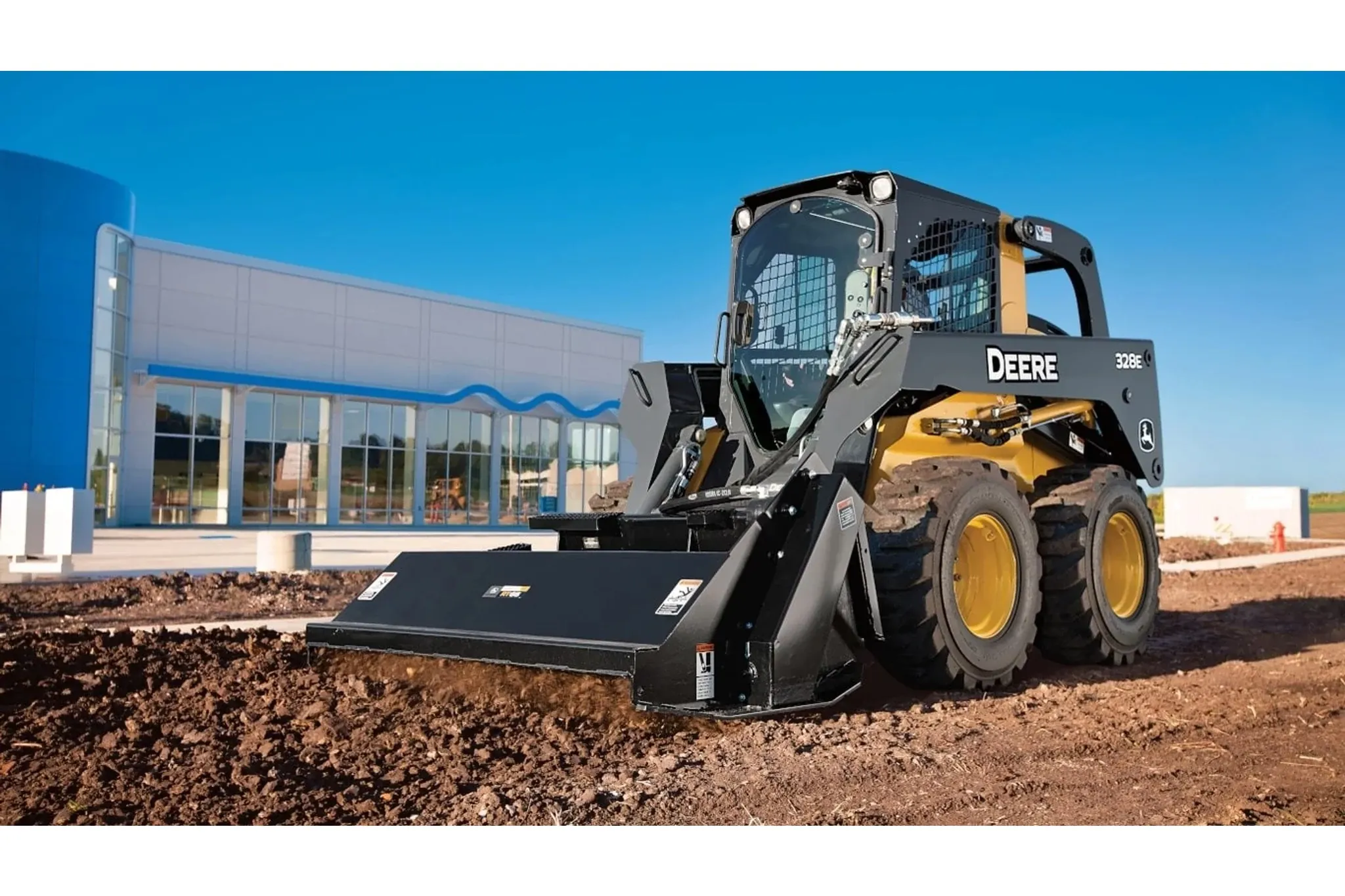 Front left view of 328E Skid Steer Loader with RT Series Rotary Tiller tilling fresh dirt in front of a large building