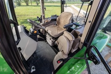 Tractor cab interior with a cushioned operator seat, a folded companion seat, control panels, steering wheel, and a view of grass and gravel path through large windows
