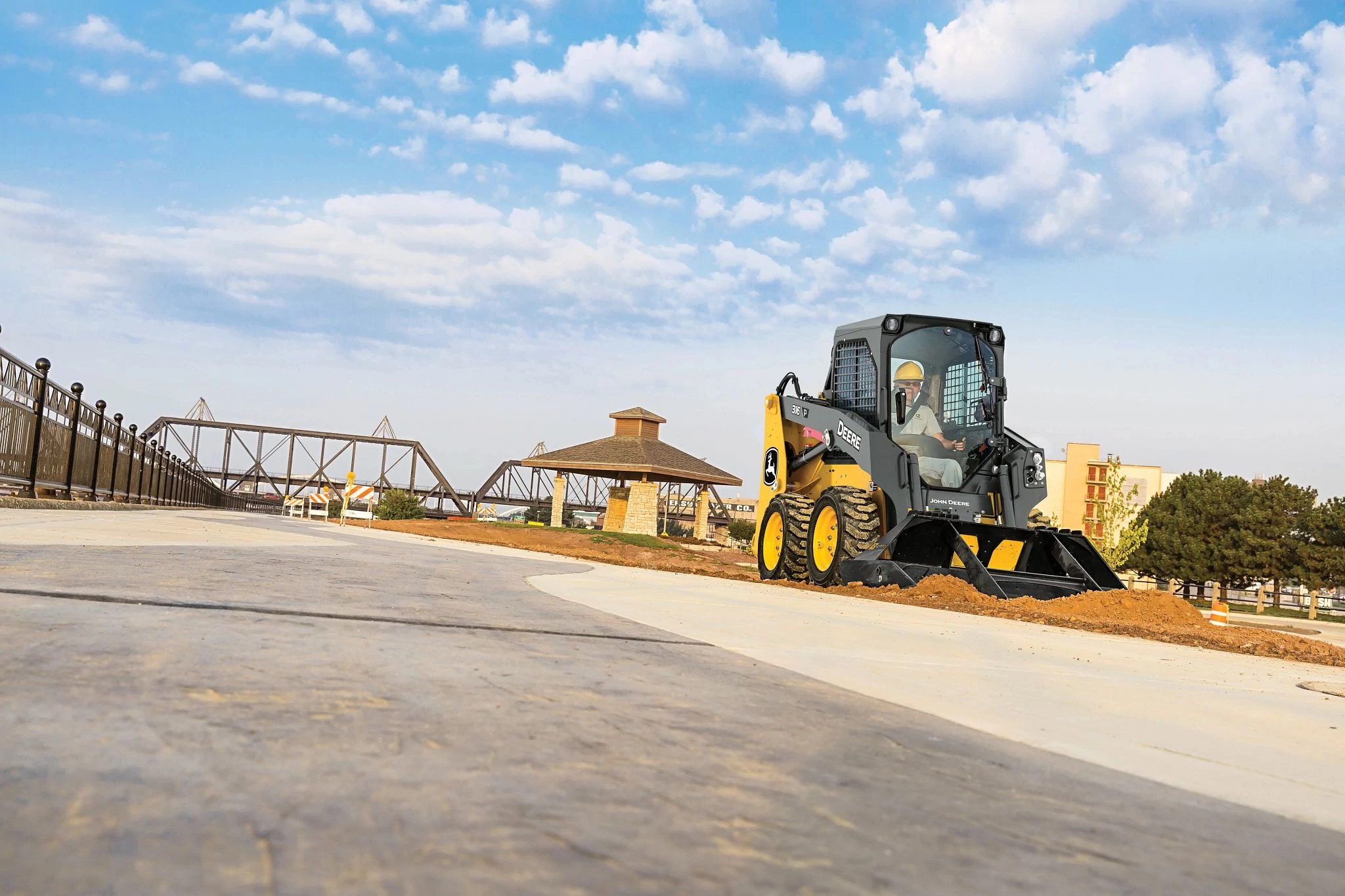 316 P-Tier skid steer loader moves along a paved surface at a construction site, pushing a small pile of reddish dirt with its front bucket.