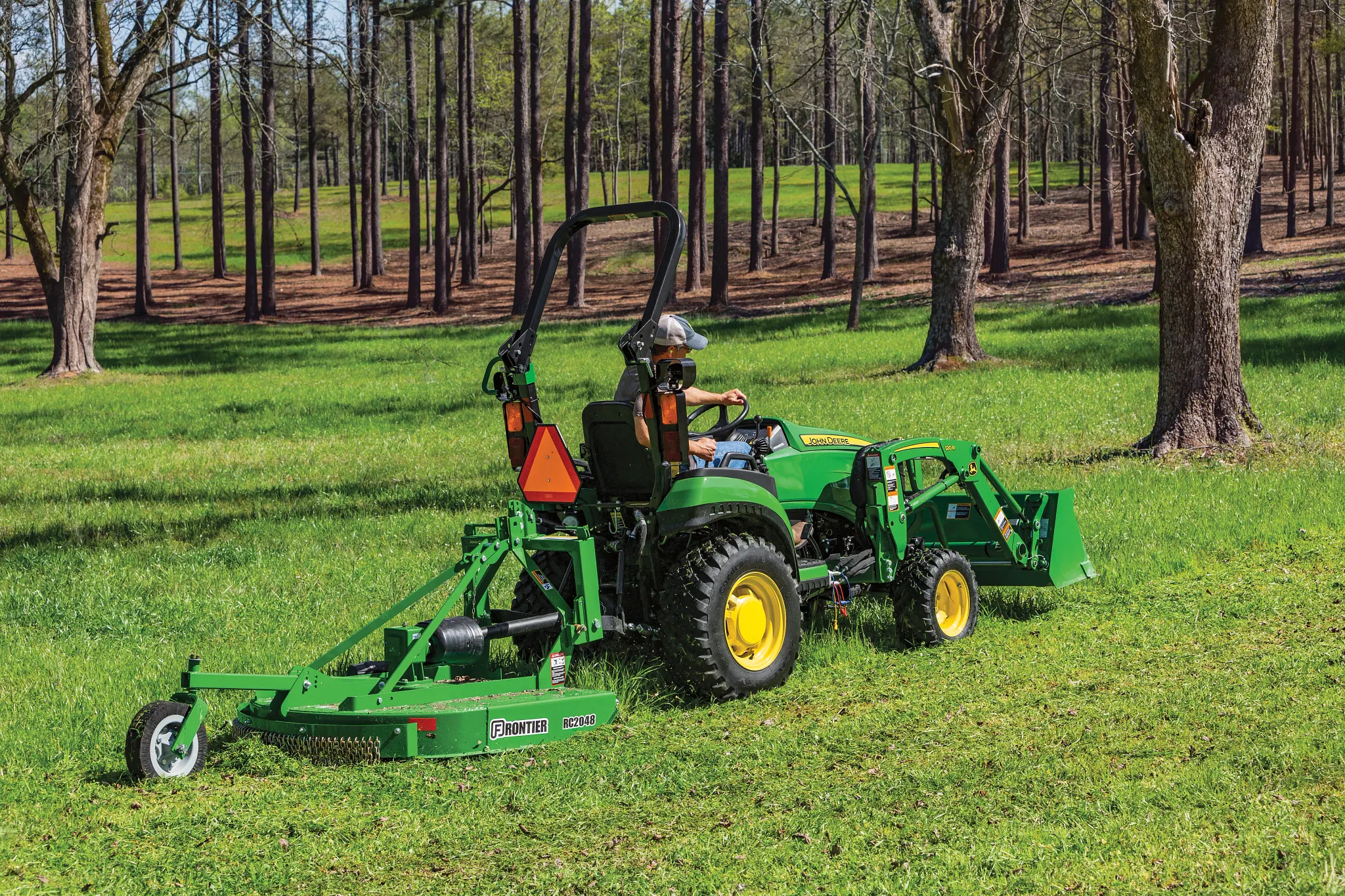 Green tractor with a front loader and rear mower attachment driving in a grassy field among evenly spaced trees in a wooded area under clear daylight