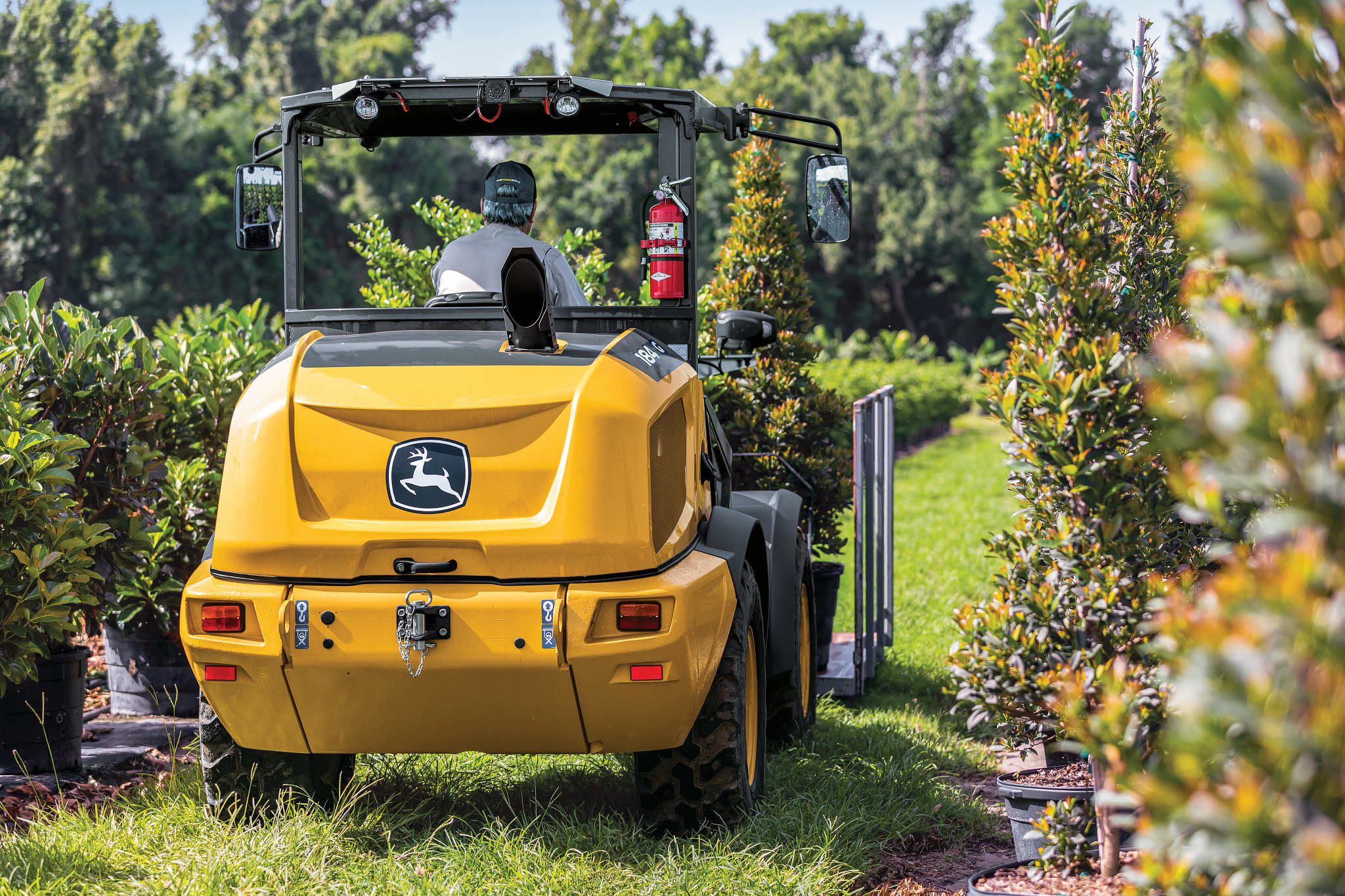Rear right view of a184 G-Tier compact wheel loader being driven by an operator past potted trees
