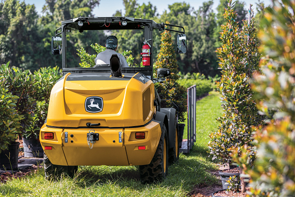 Rear right view of a184 G-Tier compact wheel loader being driven by an operator past potted trees