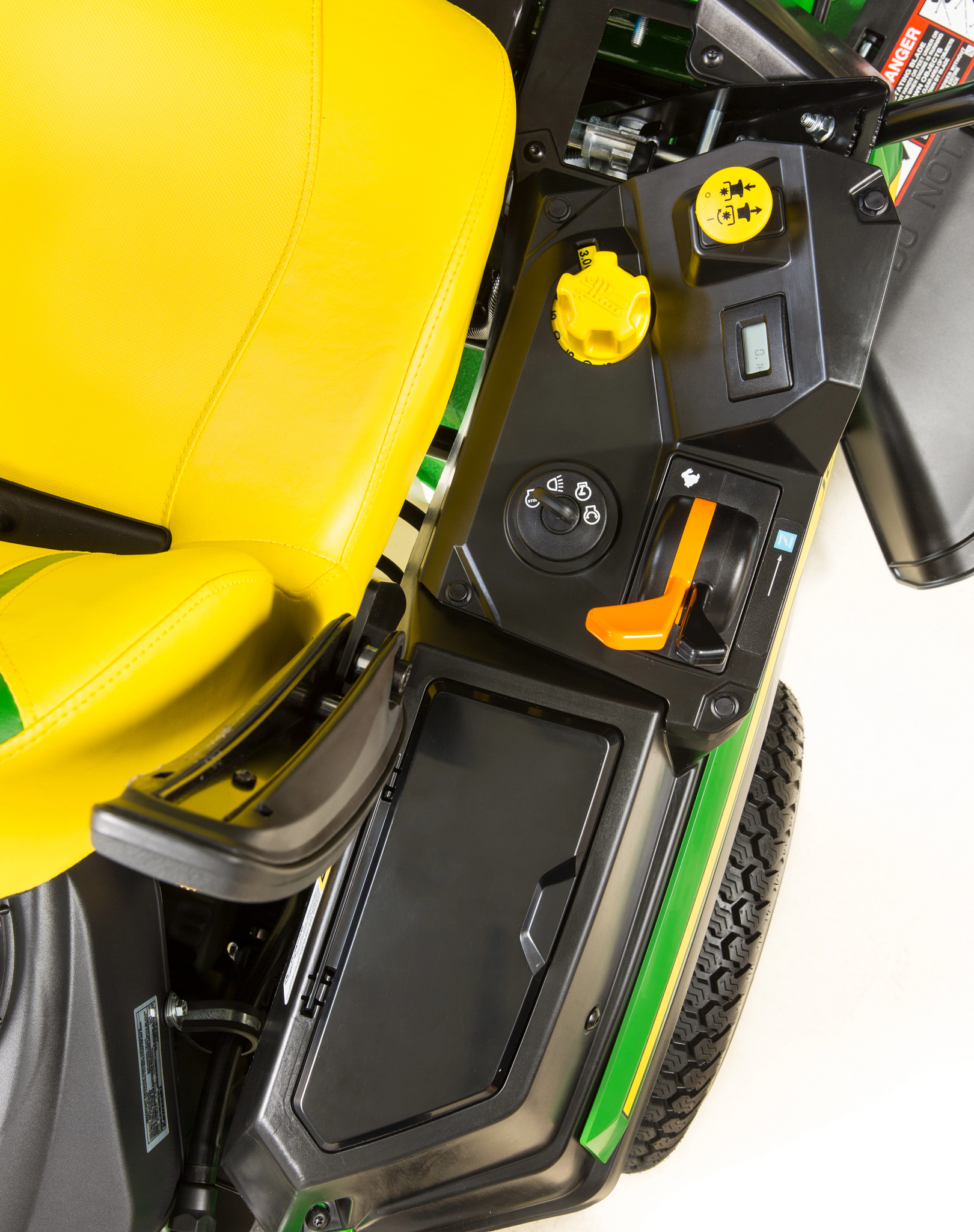 Close-up of a zero-turn mower operator area showing a yellow seat and black side control panel with a yellow height adjustment knob, ignition switch, safety icons, an orange control lever, and integrated storage compartment.