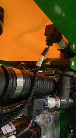 Close-up of hydraulic hoses, fittings, and an electrical sensor mounted under a planter fertilizer tank
