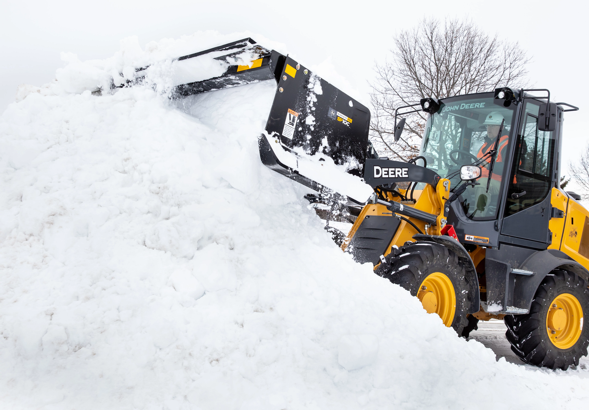 Image of a wheel loader using a front-mounted snow pusher to move a large pile of snow, with the cab, loader arms, and tires visible against a winter landscape.
