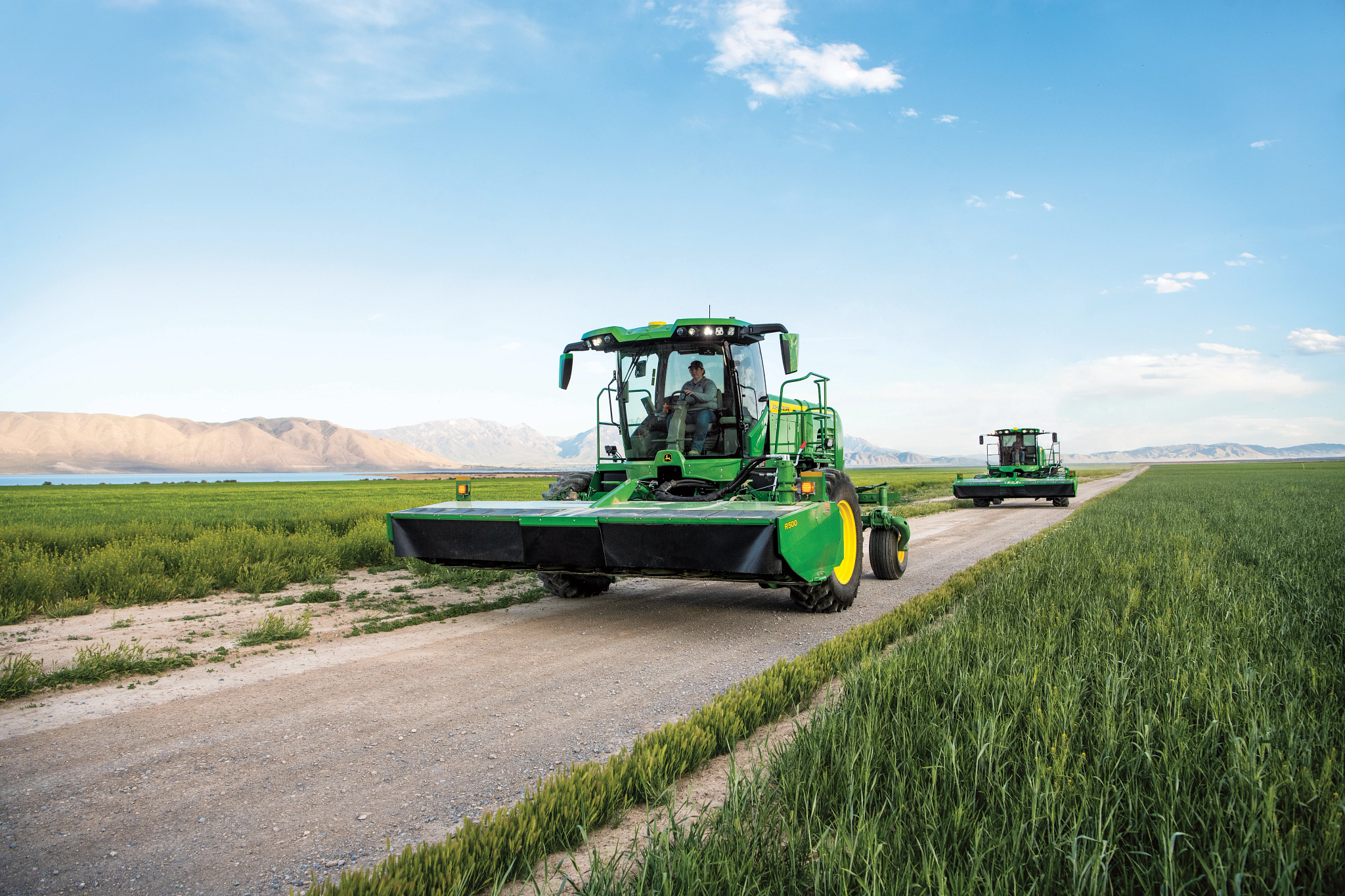 Two John Deere self‑propelled windrowers traveling in line on a dirt farm road between green fields, with wide mower‑conditioner heads raised and mountains visible in the distance under a clear sky.