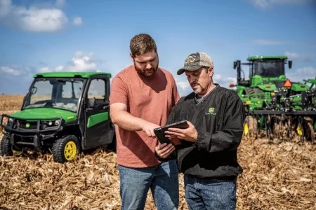 Two farmers consulting a tablet in a sunny harvested field with a Gator and tractor/coulter chisel parked on either side of them