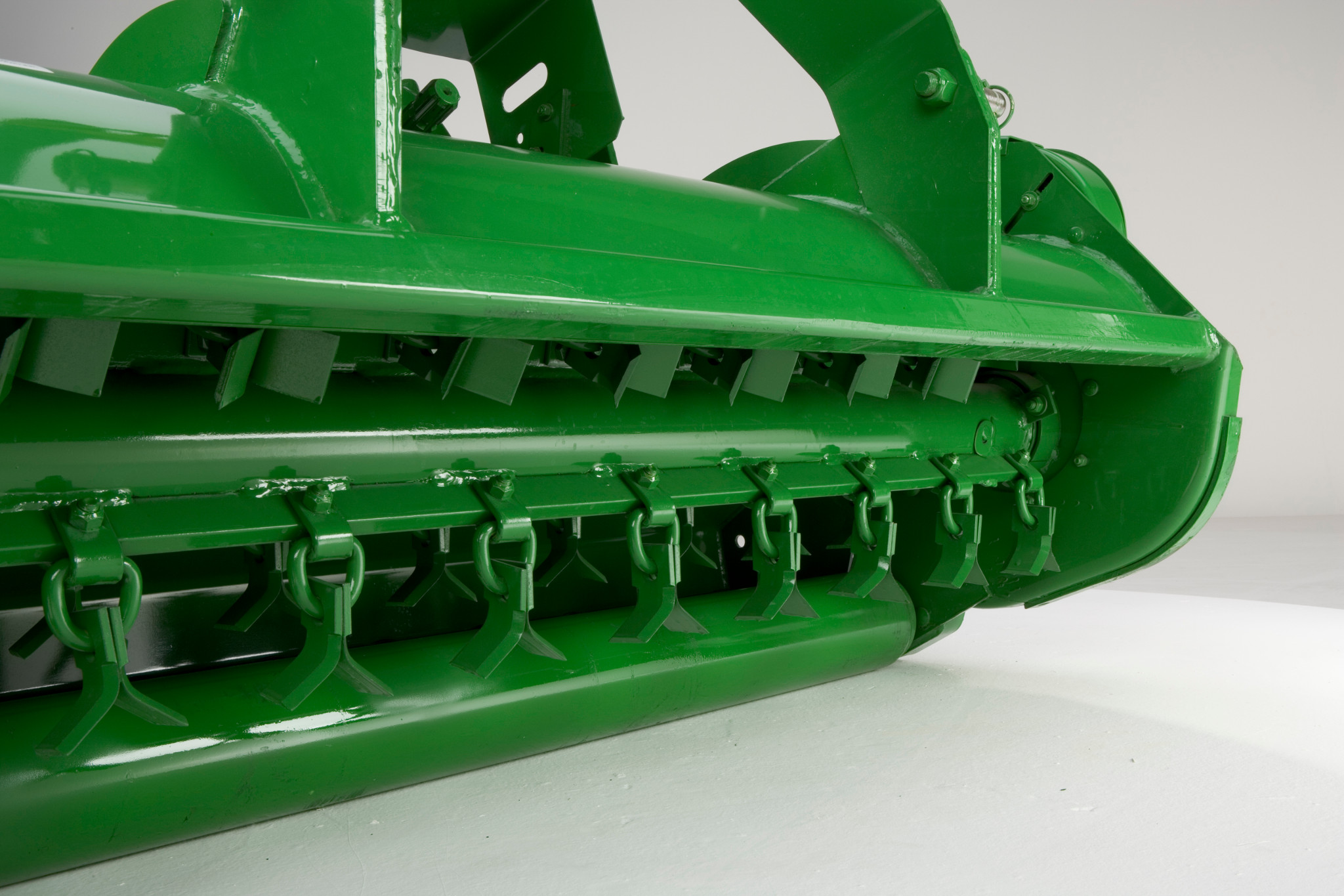Close-up of the underside of a green flail mower showing a row of hanging flail blades mounted along a rotating drum
