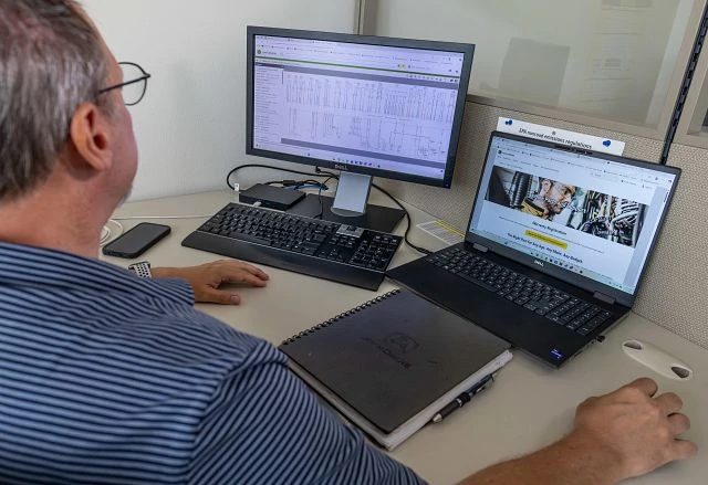 Person at a cubicle desk using a desktop monitor with a spreadsheet and a laptop displaying a website, with a keyboard, mouse, smartphone, and closed notebook on the desk