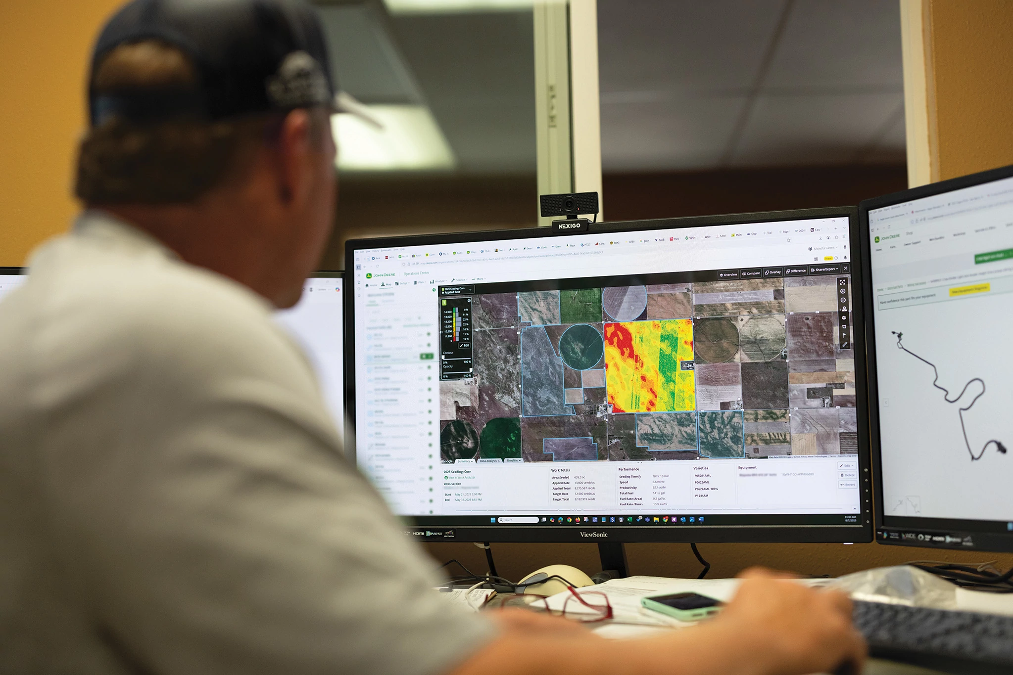A person at a desk reviews field performance and agronomic data with maps and machine activity displayed across multiple computer monitors.