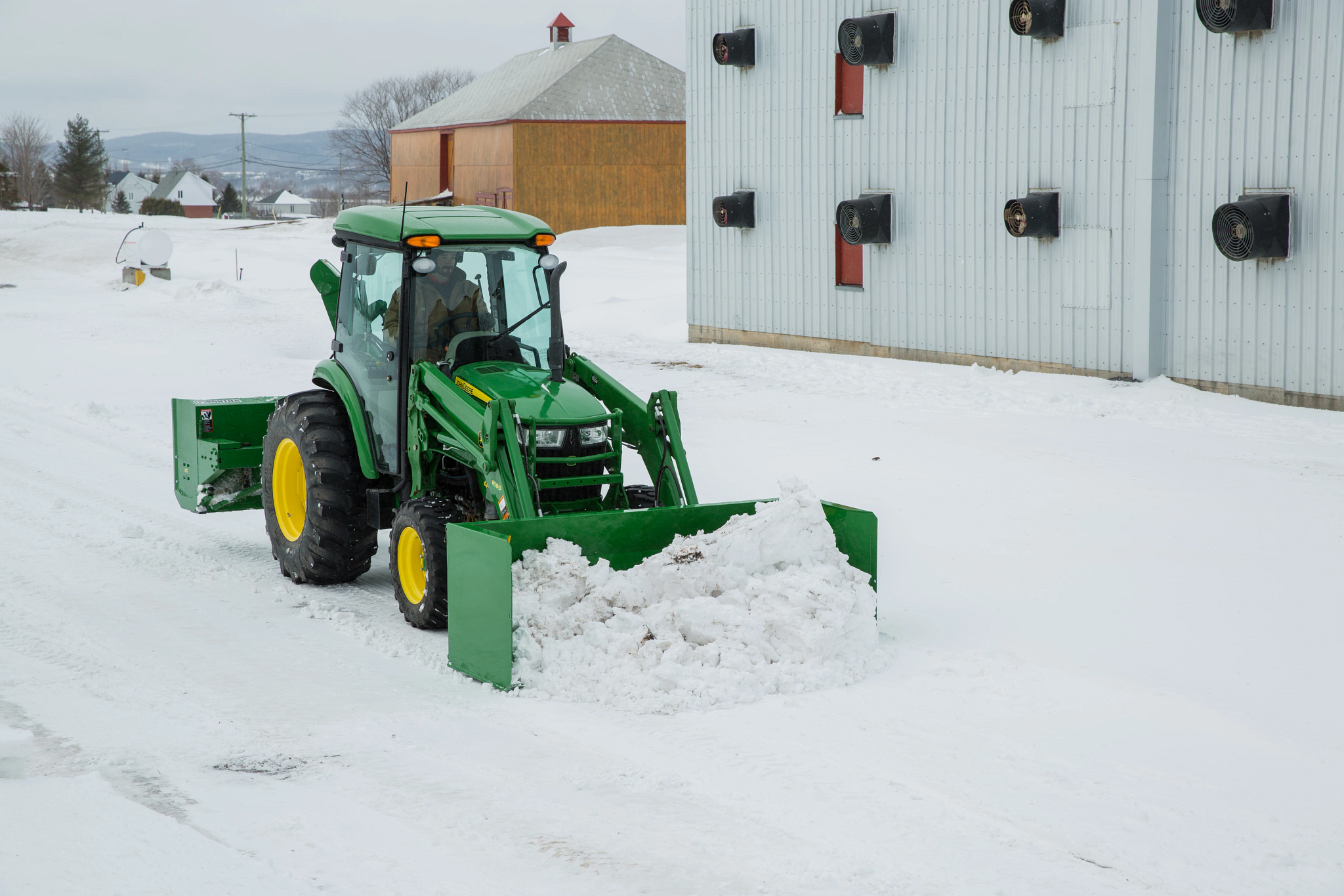 Front right view of a John Deere 4-Series tractor handling snow with the AS10H Snow Push beside two outbuildings