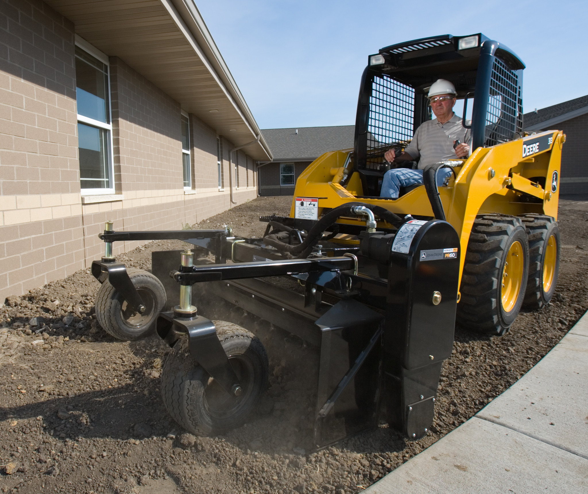 Front left view of angle power rake mounted on front of a skid steer loader raking dirt along a sidewalk next to a building