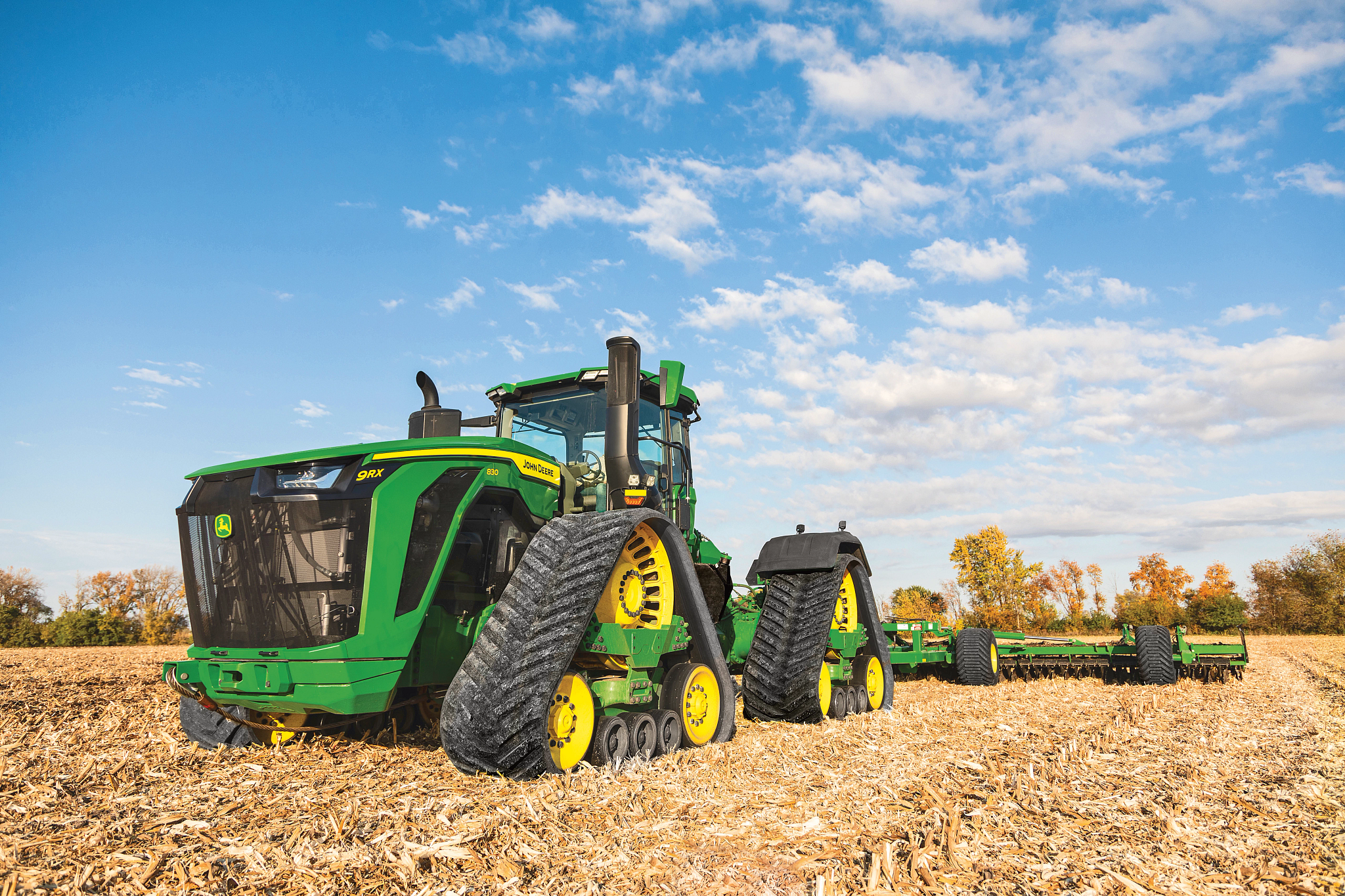 Large green tracked tractor in a harvested field covered in crop residue with a matching green implement trailing behind and a blue sky with scattered clouds and trees lines the horizon in the background