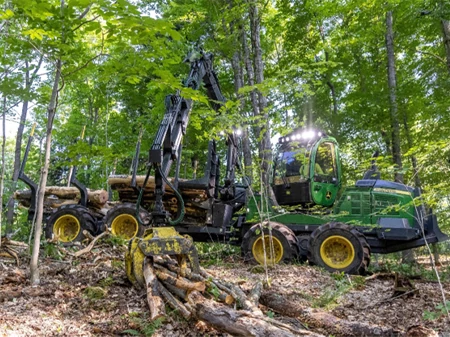Side-right view of a 1110G forwarder picking up fallen logs in a forest to add to its half-full open bed