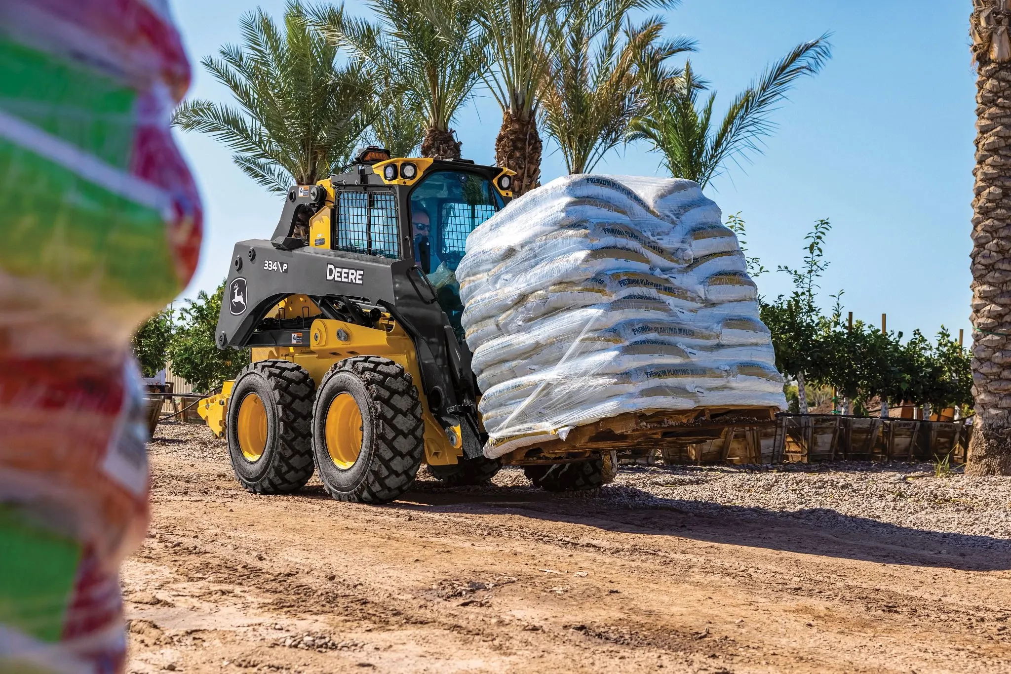 334 P-Tier skid steer loader transports a large pallet stacked with white bagged materials along a dirt path.