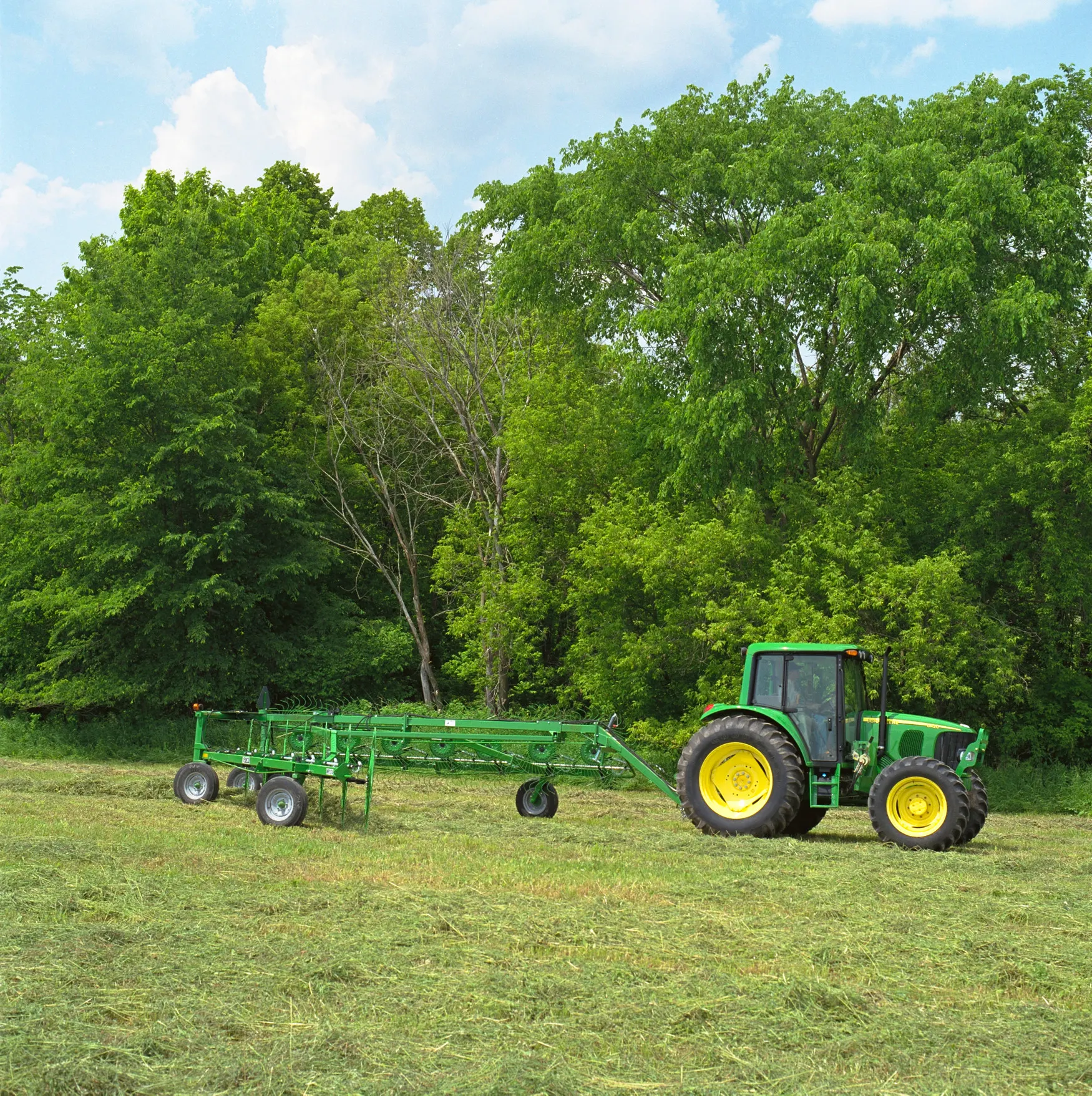 Side right view of a tractor using a green WR High-Capacity Wheel Rake in a hay field bordered by trees