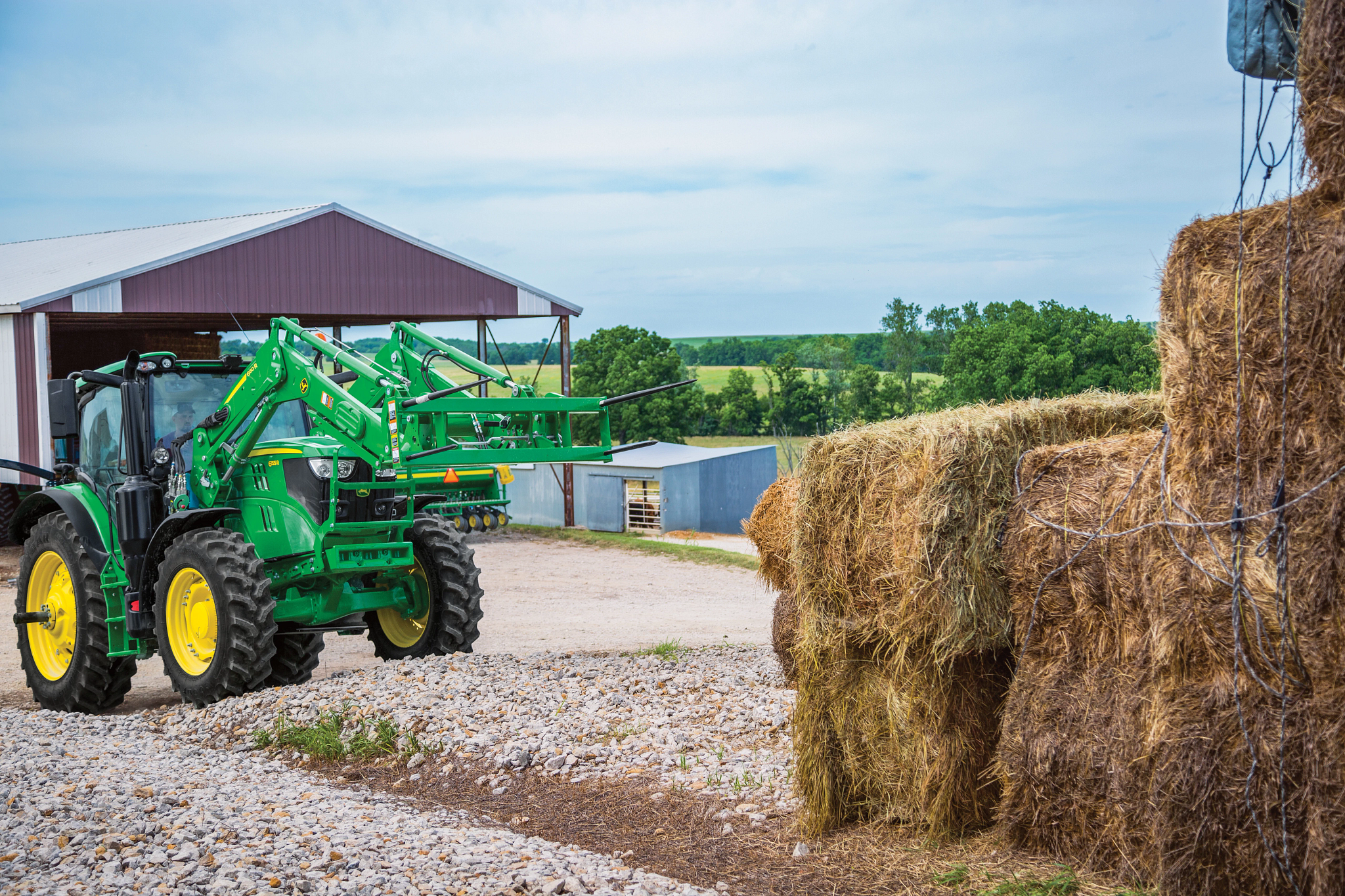 A tractor using a bale spear to lift hay bales on a gravel drive