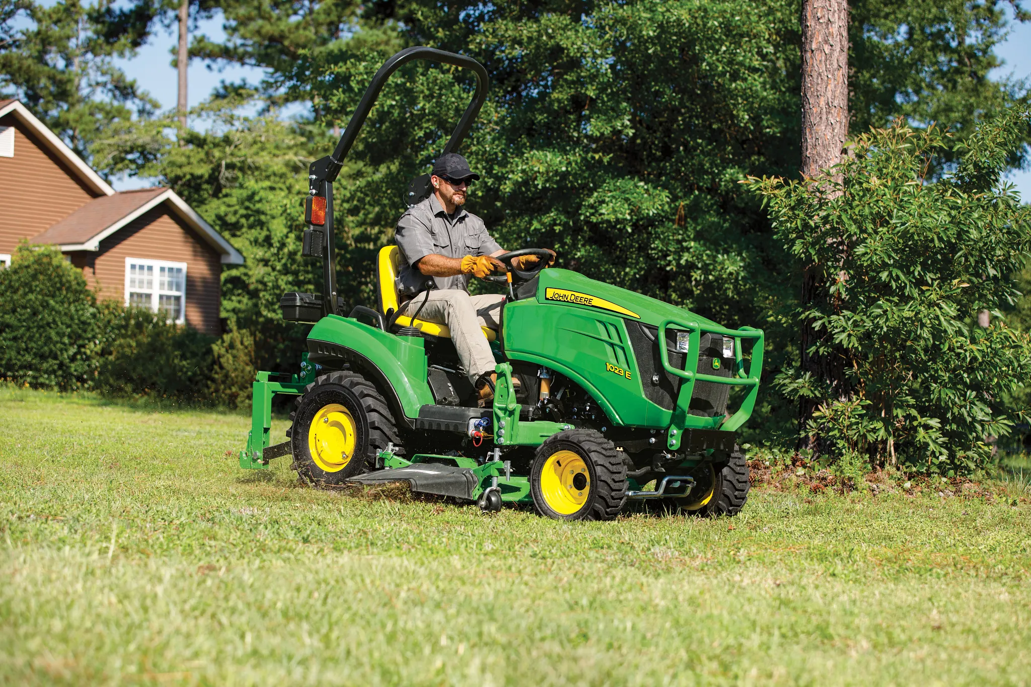 Person operating a green compact tractor with a mower attachment cutting grass across a yard near a house, with trees and shrubs in the background