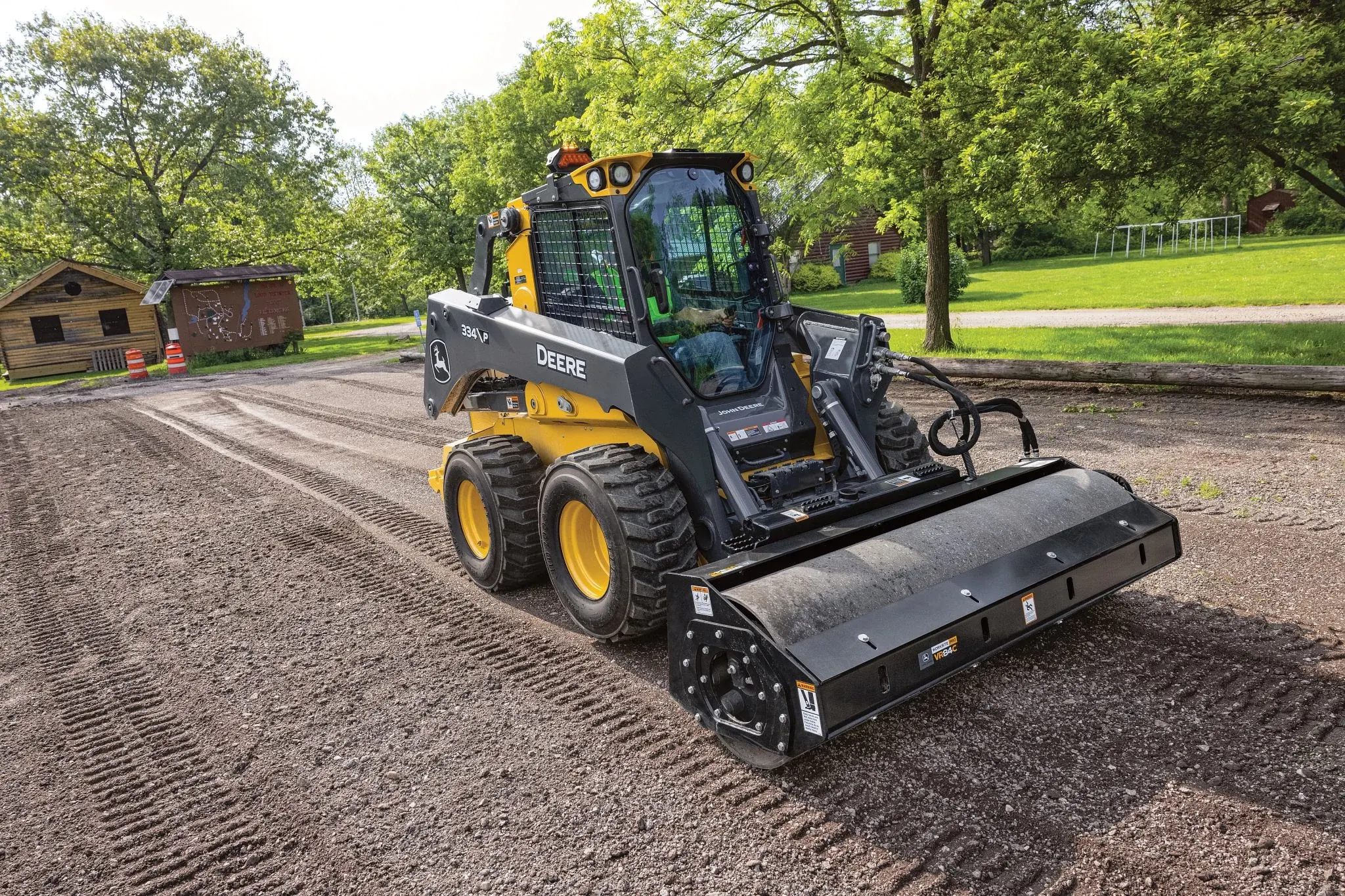 334 P-Tier skid steer loader equipped with a large cylindrical brush attachment cleans and smooths a dirt surface in a park-like setting.
