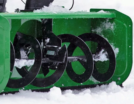 Close-up of a snow blower housing with rotating black auger blades partially covered in snow, actively pulling and breaking up snow.