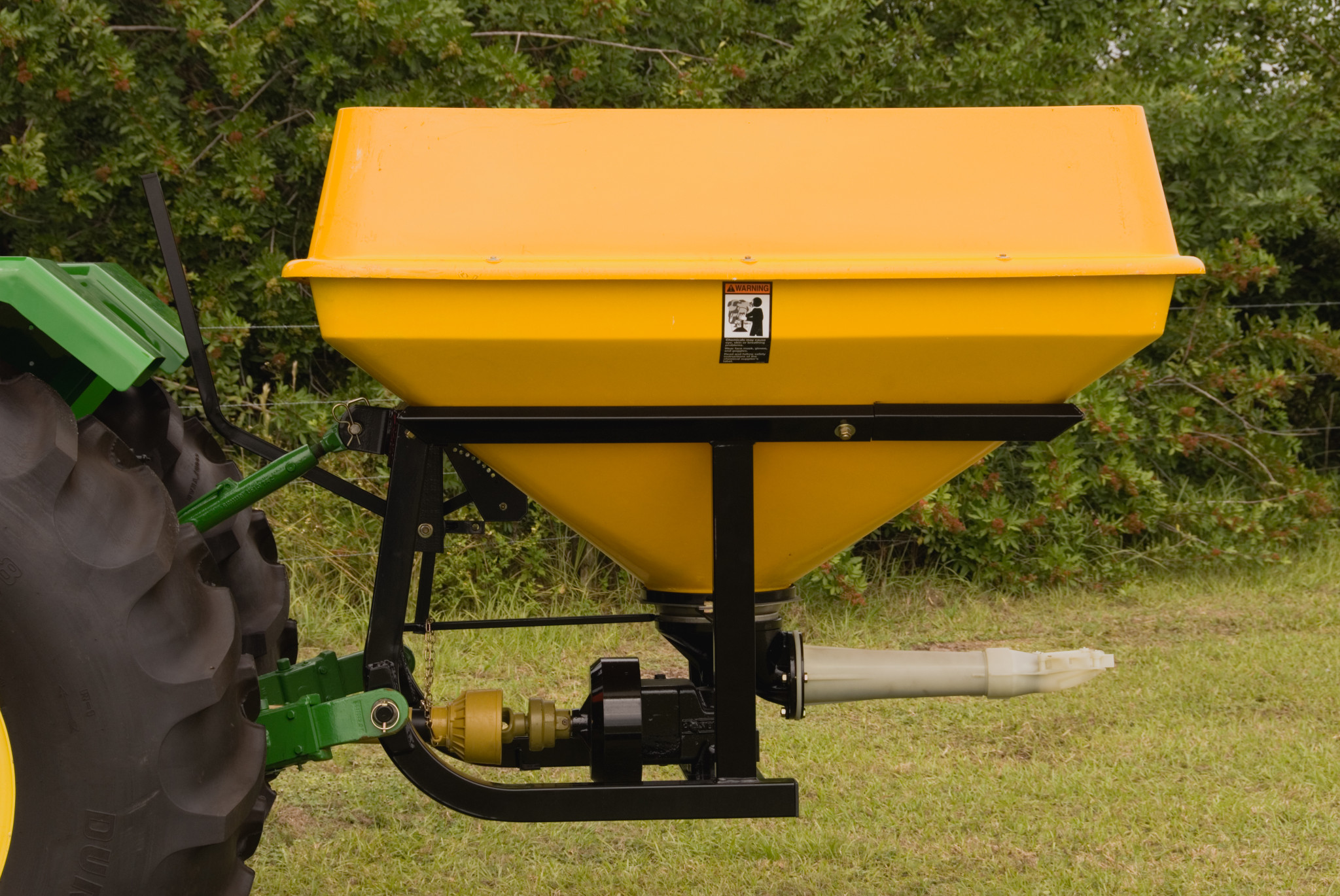 Side view of a yellow cone-shaped pendular spreader mounted on the rear of a tractor, positioned on grass with trees in the background