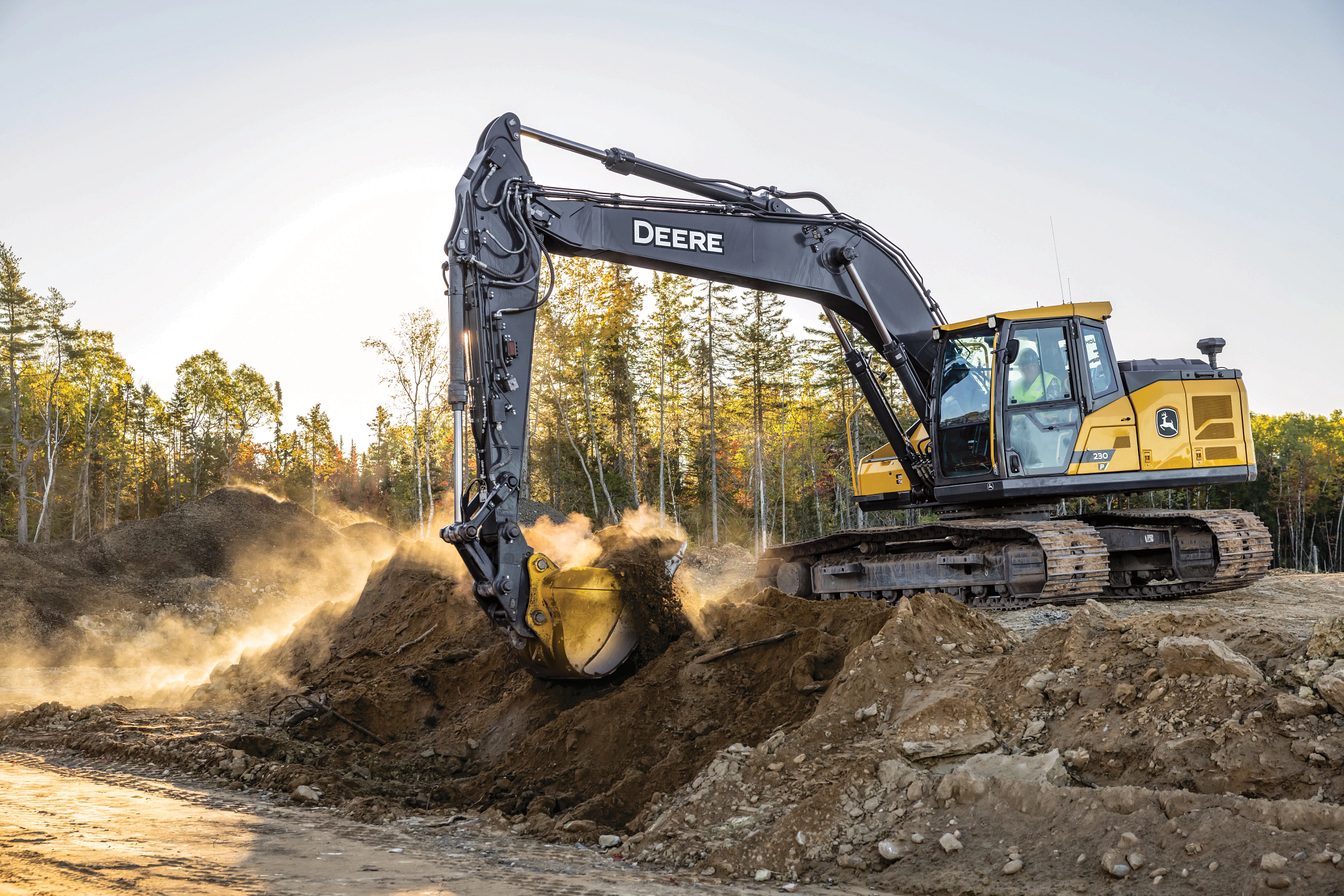 Side left view of a 230 P-Tier excavator scooping dirt from atop a pile on a tree-lined jobsite