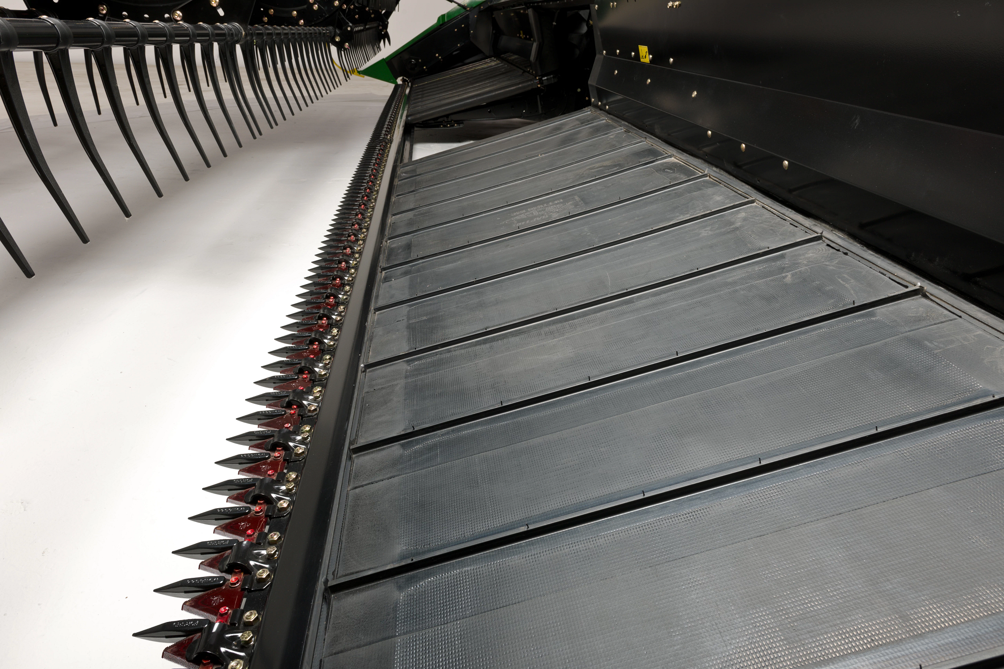 Close-up of a windrower header showing the cutterbar with sharp triangular knives and the belt behind it, with pickup tines visible at the top edge