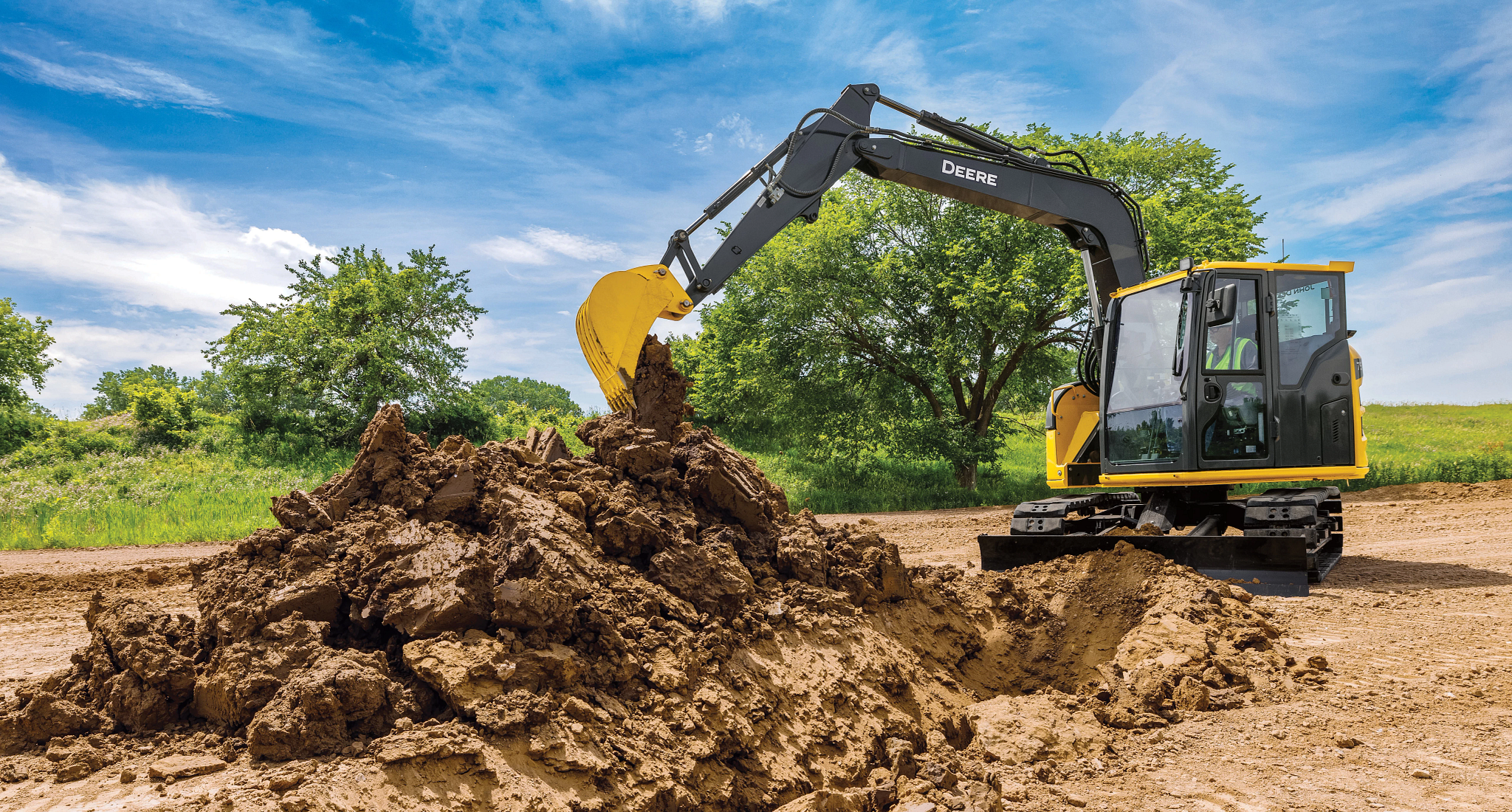 Yellow John Deere excavator with a black arm and bucket digging into a large pile of brown soil at a construction site