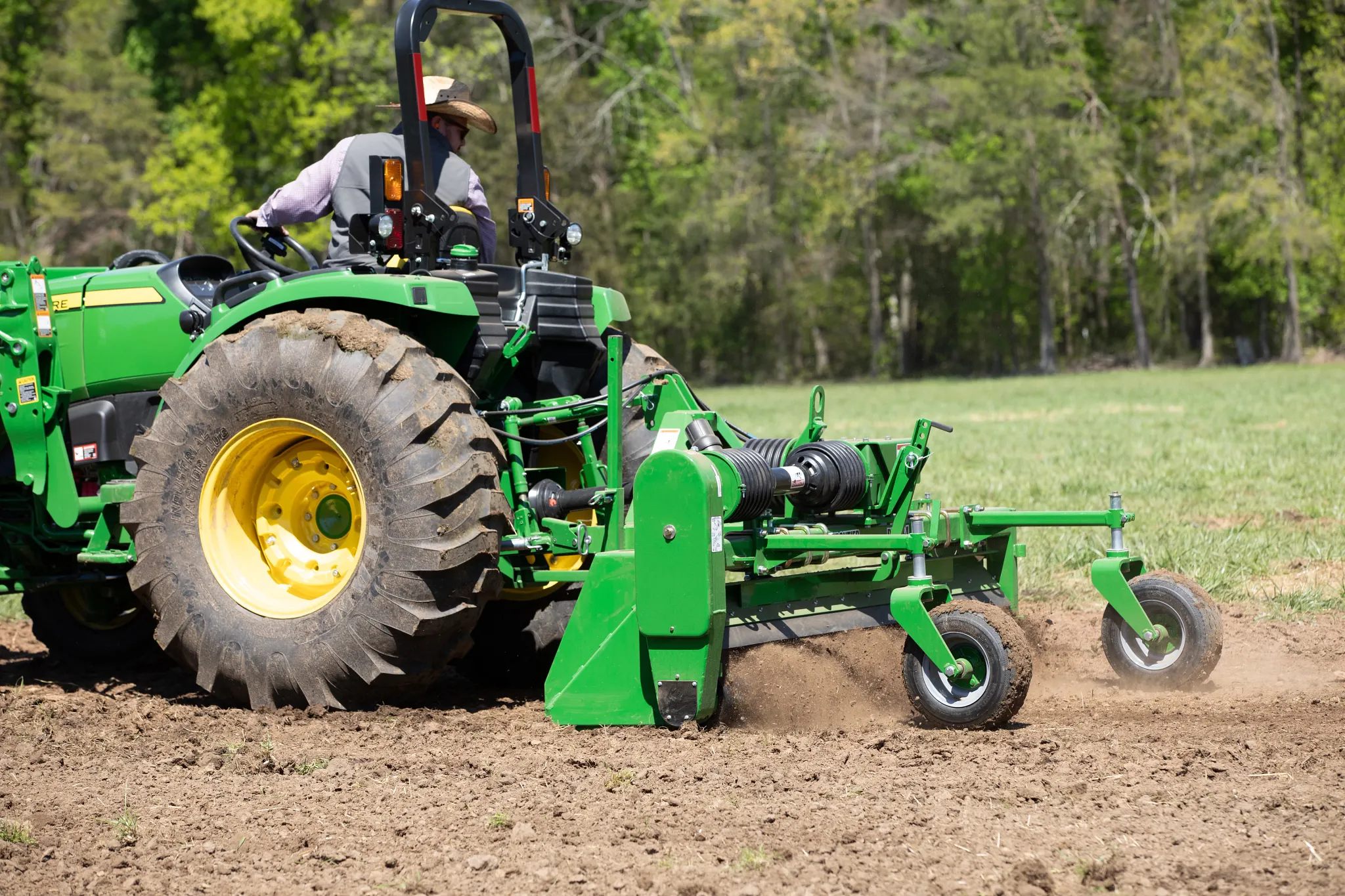 Tractor tilling a field, with soil turning behind the implement and trees in the background.