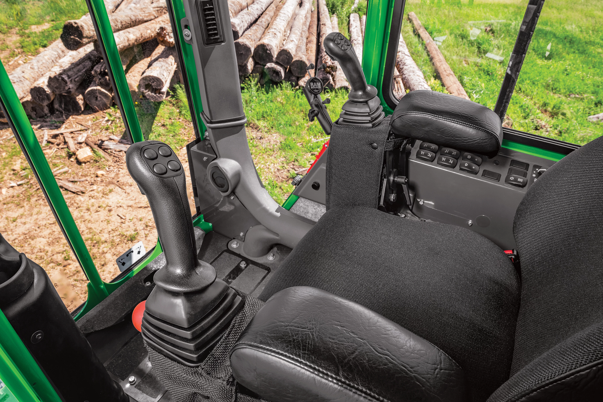 Interior of a knuckleboom loader cab showing a padded seat with armrest buttons, and dual joysticks, with stacked logs visible outside the window
