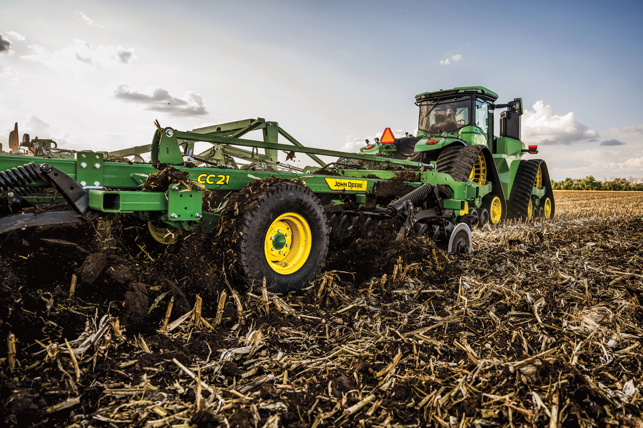 Green tractor pulling a wide tillage implement across a harvested field, churning soil and crop residue under a partly cloudy sky in a rural farmland setting with corn residue