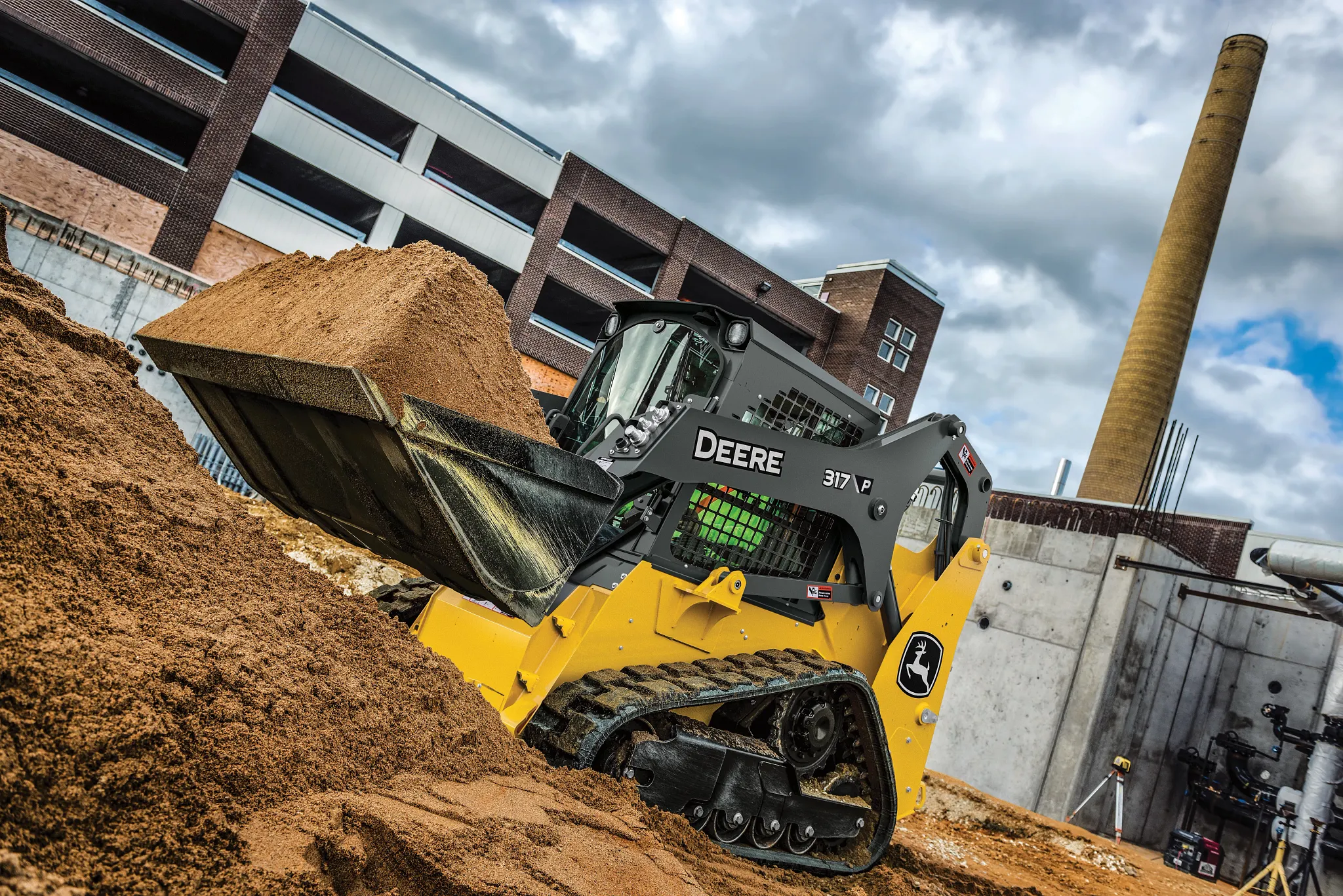 John Deere 317 compact track loader with a black front bucket, gray and yellow body, enclosed operator cab protected by metal mesh, and rubber tracks, positioned on a plain light gray background.
