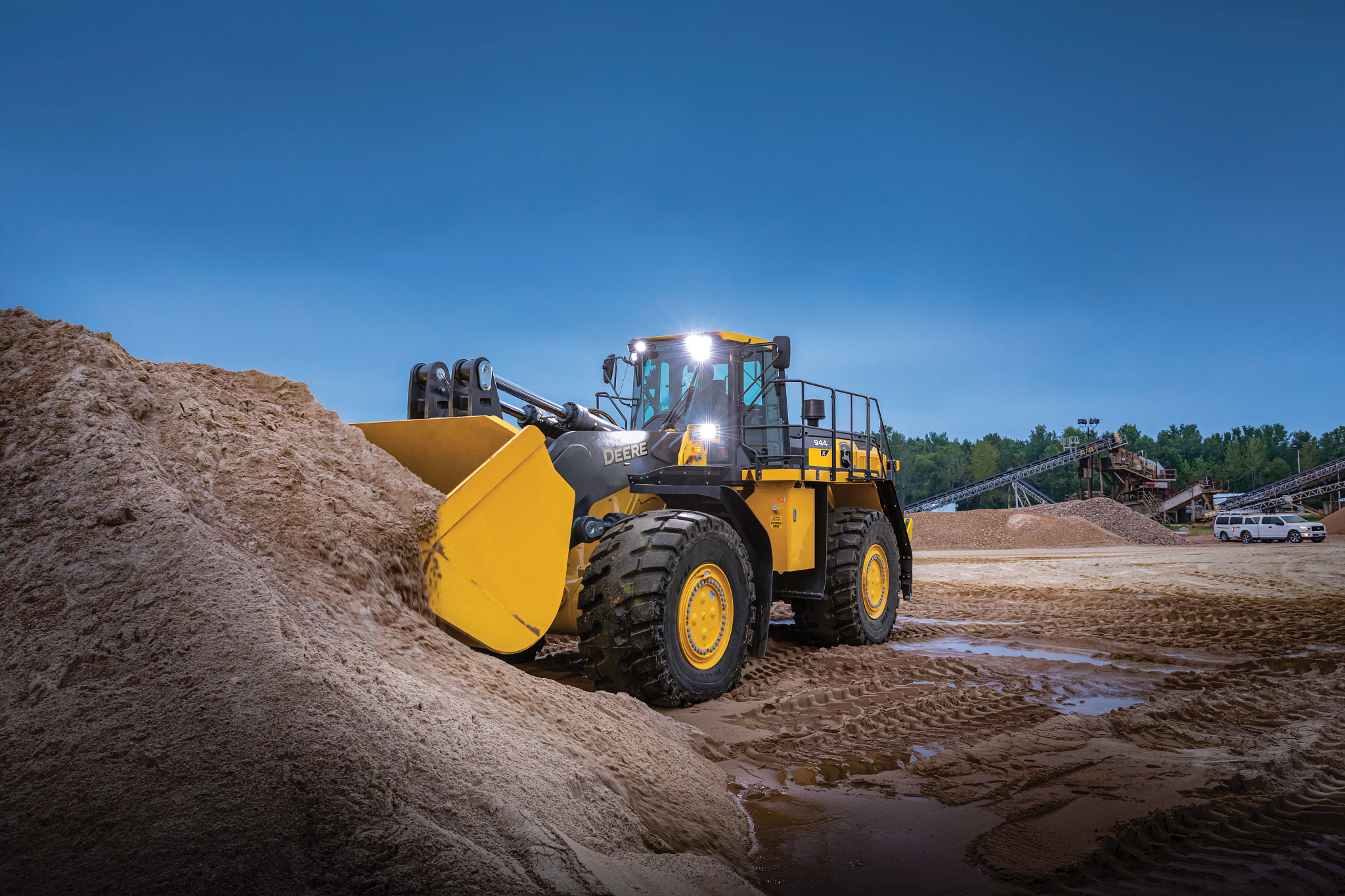 A large yellow wheel loader with bright headlights pushes into a mound of sand at a quarry