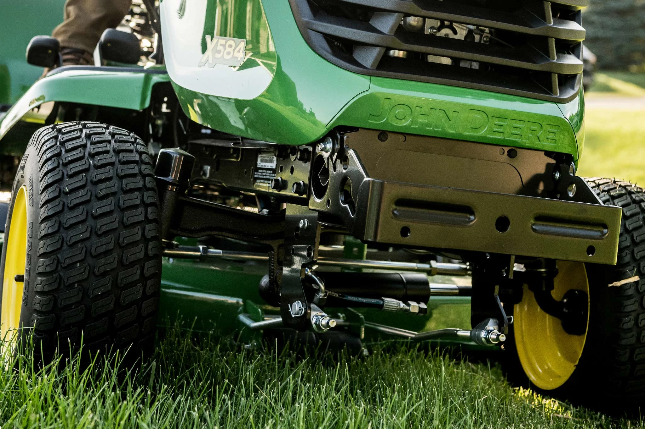 A low-angle view of a John Deere riding lawn mower showing the front tires, yellow rims, and visible steering and suspension components as it sits on green grass