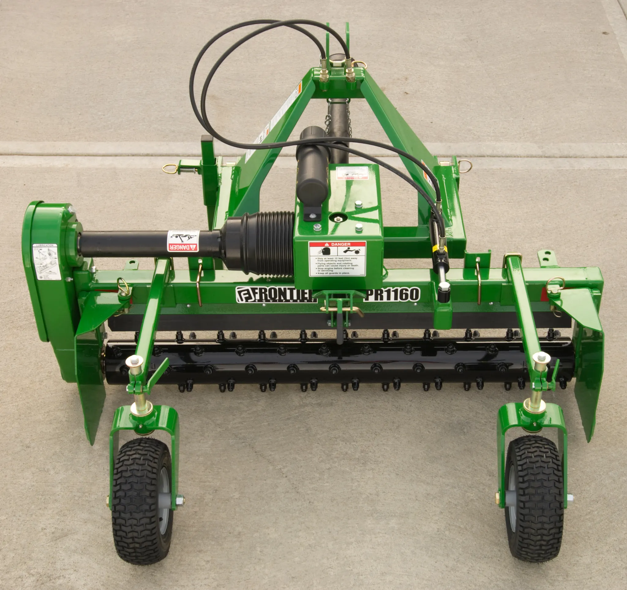 Front view of a green Frontier PR1160 agricultural implement with a steel frame, hydraulic hoses, rotating cutting drum, adjustable side skids, and two black rubber support wheels on a concrete surface.