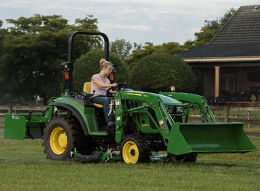 Woman operating a green compact tractor with a front-end loader and mower deck, driving across a grassy lawn near a house and trees on a clear day