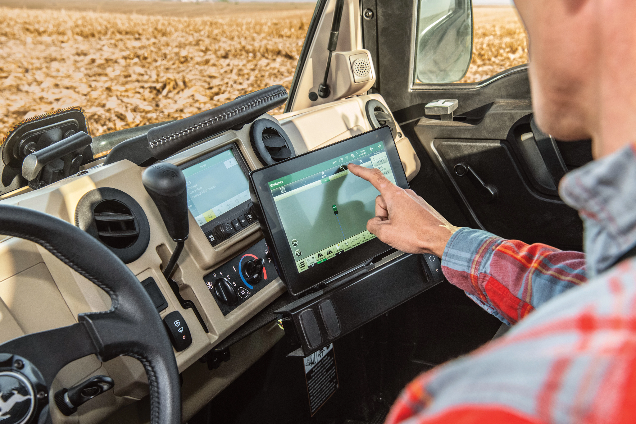 Over-the-shoulder view of an operator touching a button on a Universal Display screen within the cab of an XUV845R Gator. A golden, harvested field can be seen through the windows