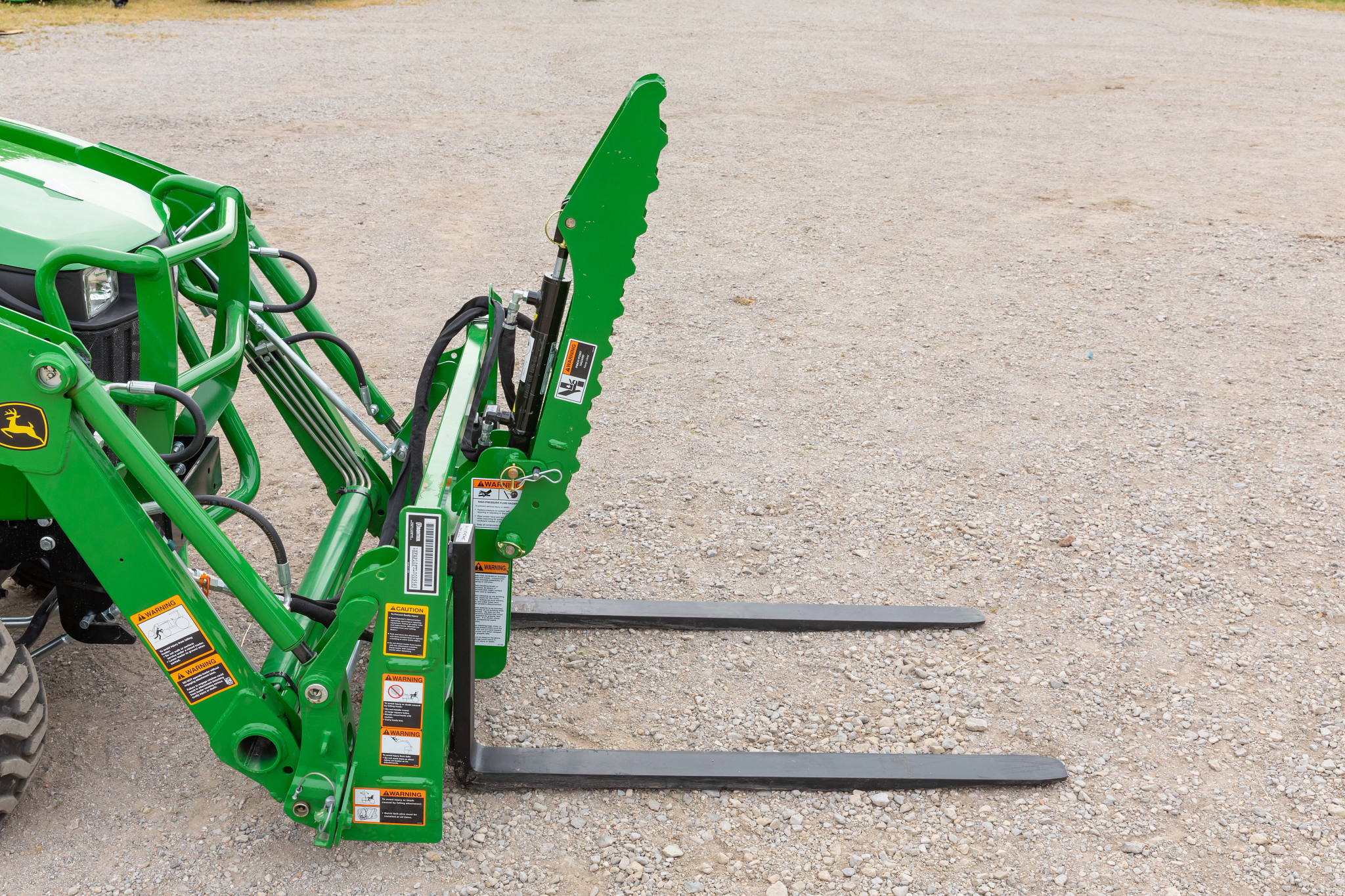 Tractor front-loader attachment with pallet forks and a vertical serrated grapple arm positioned on a gravel surface.