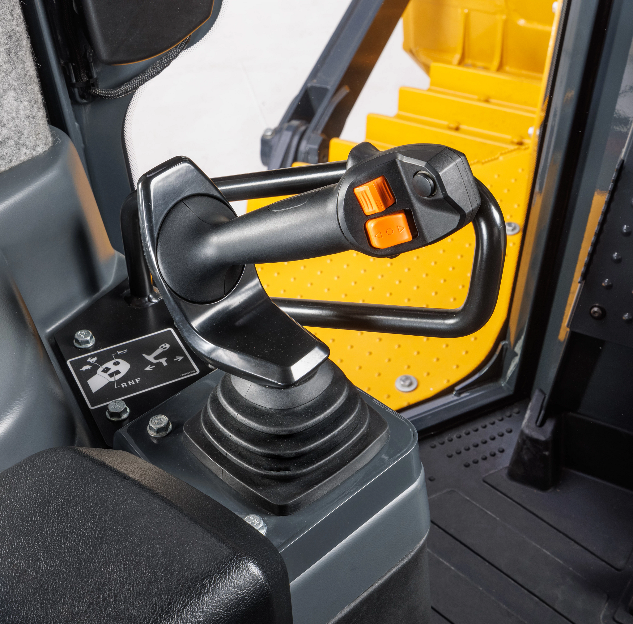 Close-up of a control joystick inside heavy equipment, featuring a black handle with orange buttons mounted on a console, with yellow perforated metal steps and interior cab panels visible in the background.