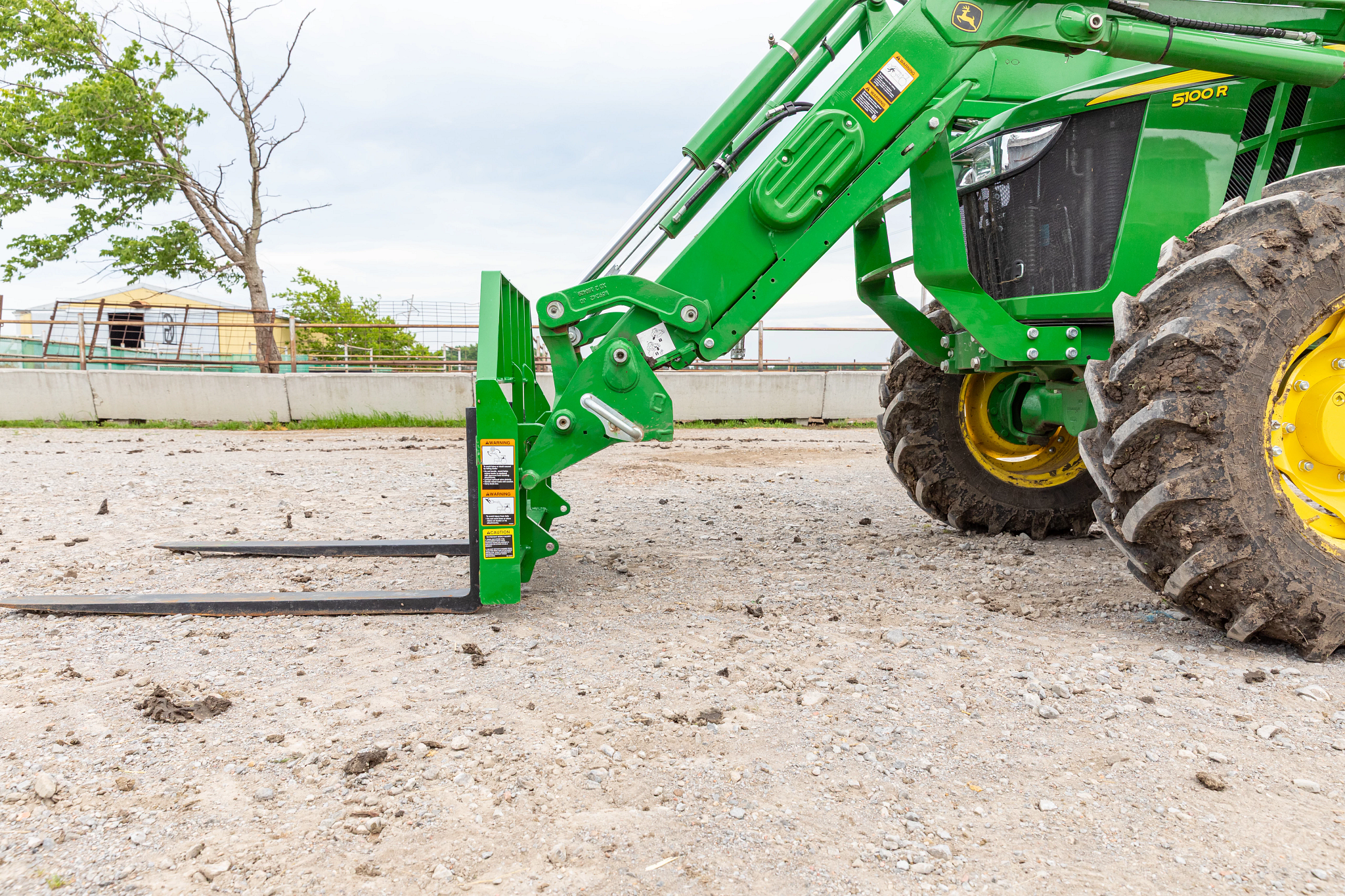 Side view of tractor with a front‑mounted pallet fork attachment lowered to the ground, showing the loader arms, fork tines, large rear tire, and model marking “5100R”.