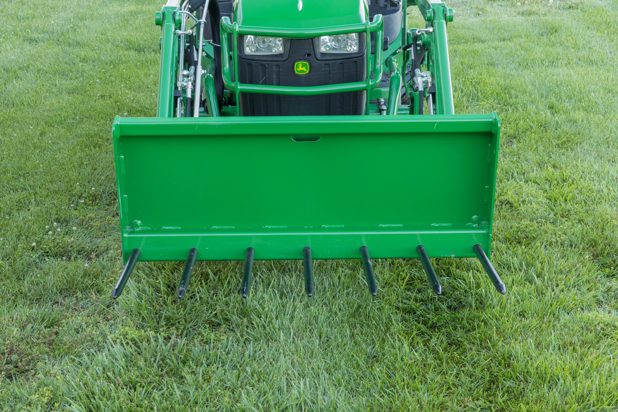 Front view of tractor equipped with a front-mounted bucket or grapple attachment with evenly spaced metal tines with loader arms visible behind the attachment.